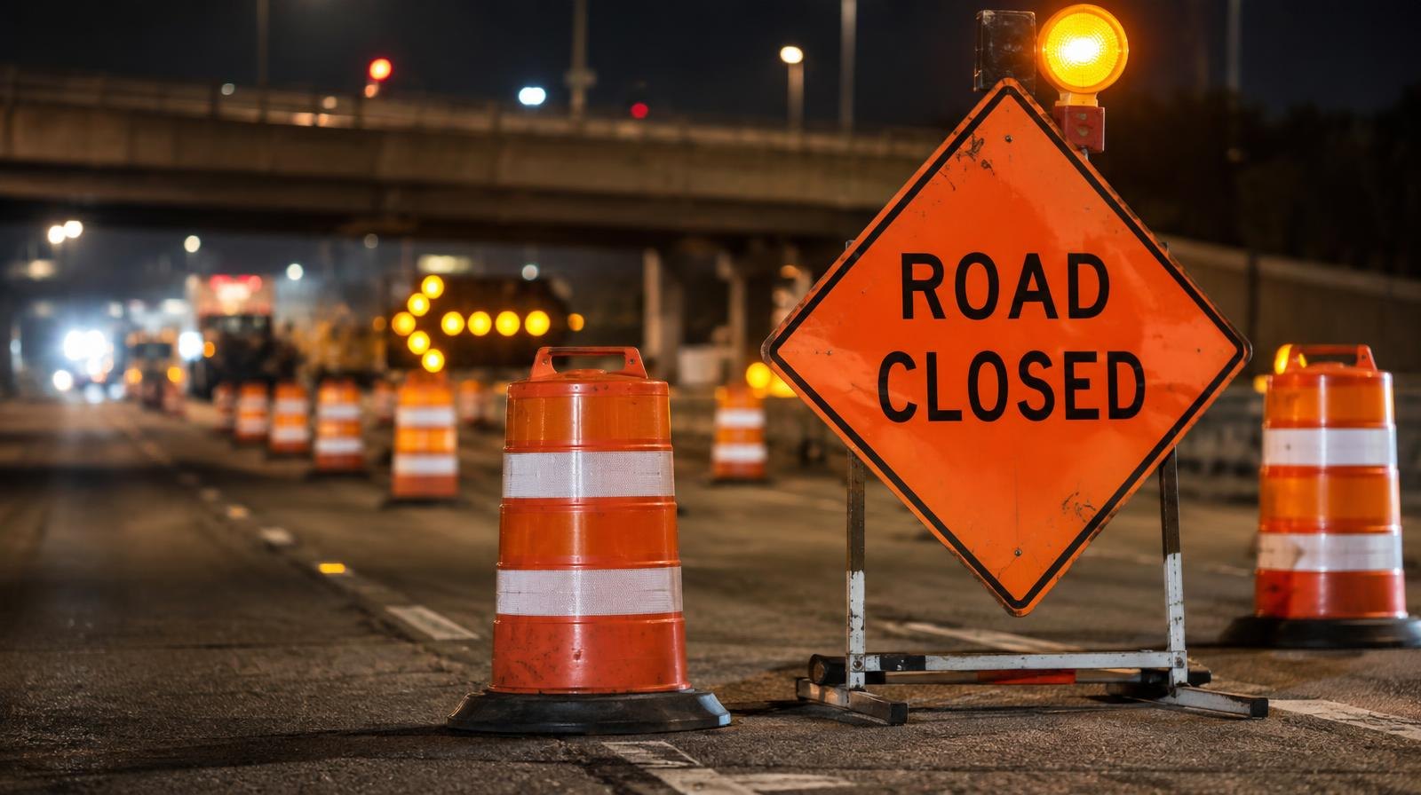 Orange road closure signs and traffic cones blocking lanes on a highway at night.