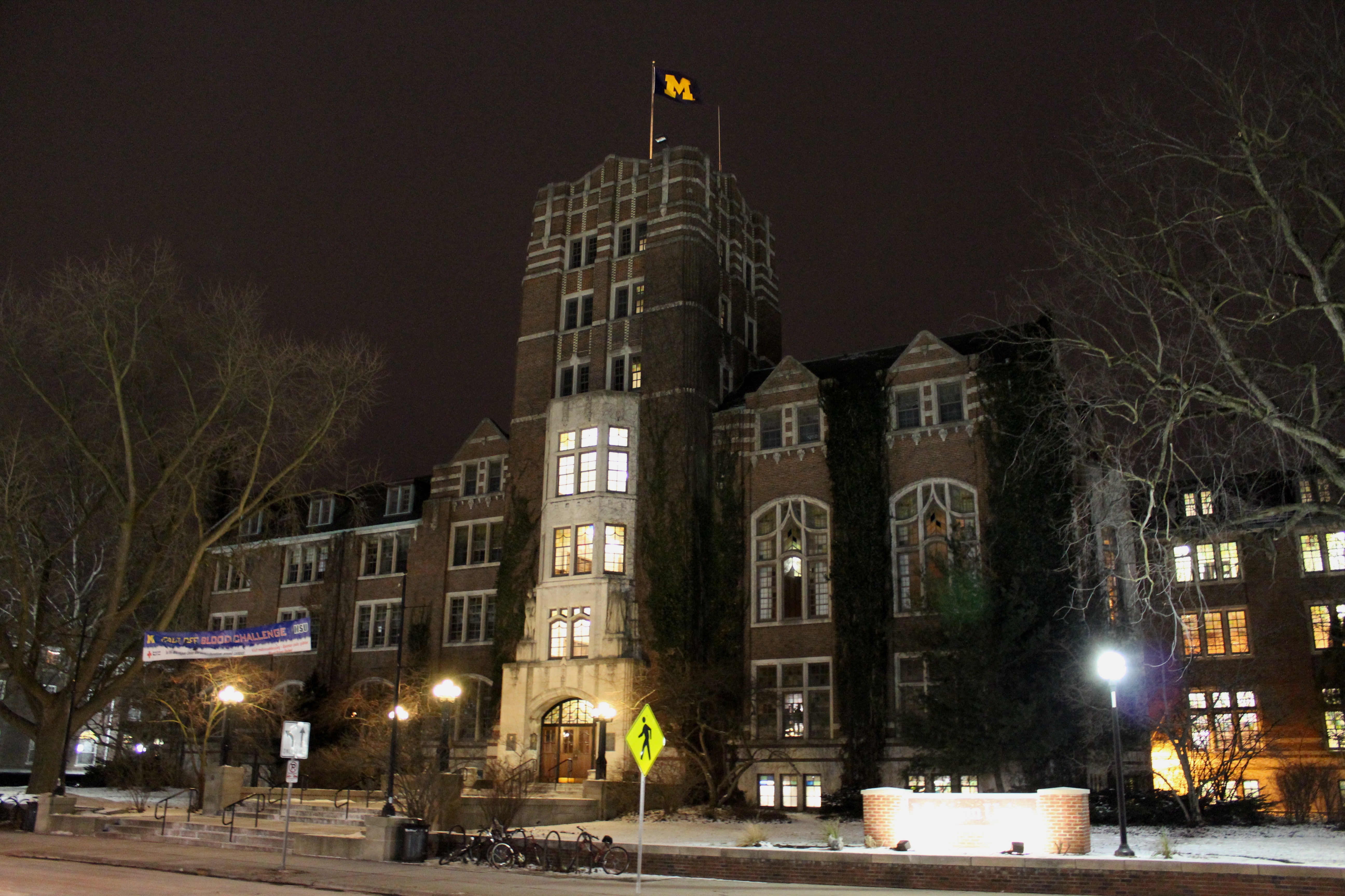 Gothic brick university building lit at night in winter with a flag atop its central tower.