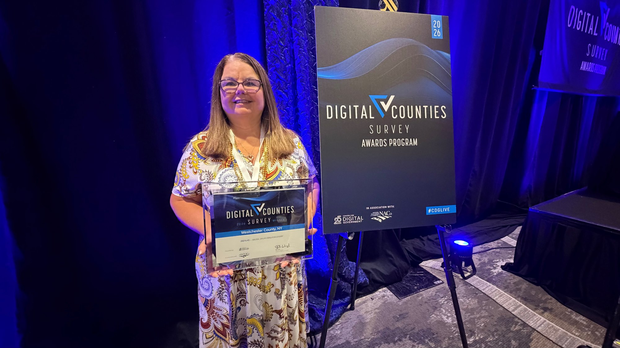 Woman in floral dress holding a Digital Counties Survey award certificate at a conference.