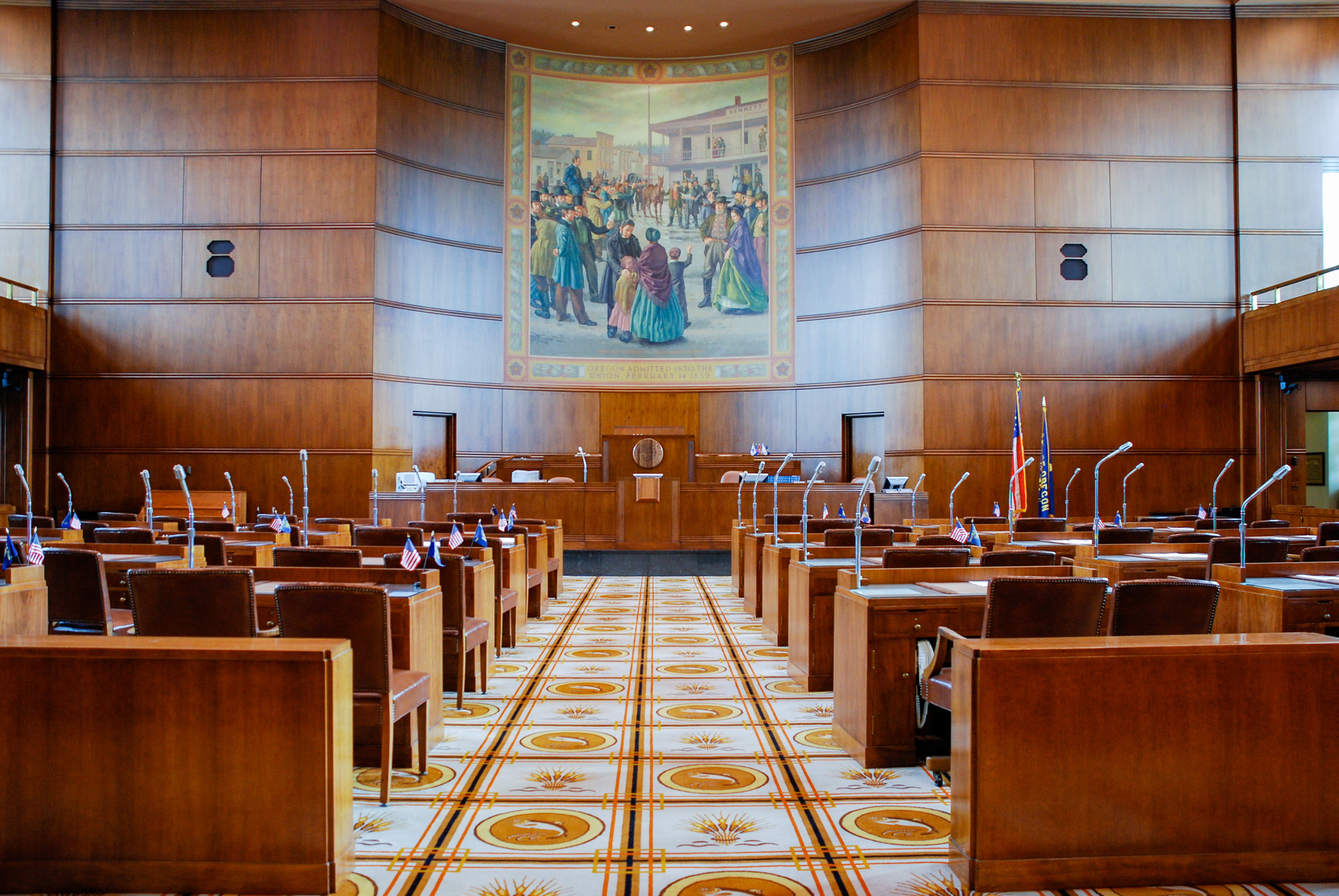 Empty legislative chamber with wooden desks, microphones, and a large historical mural.