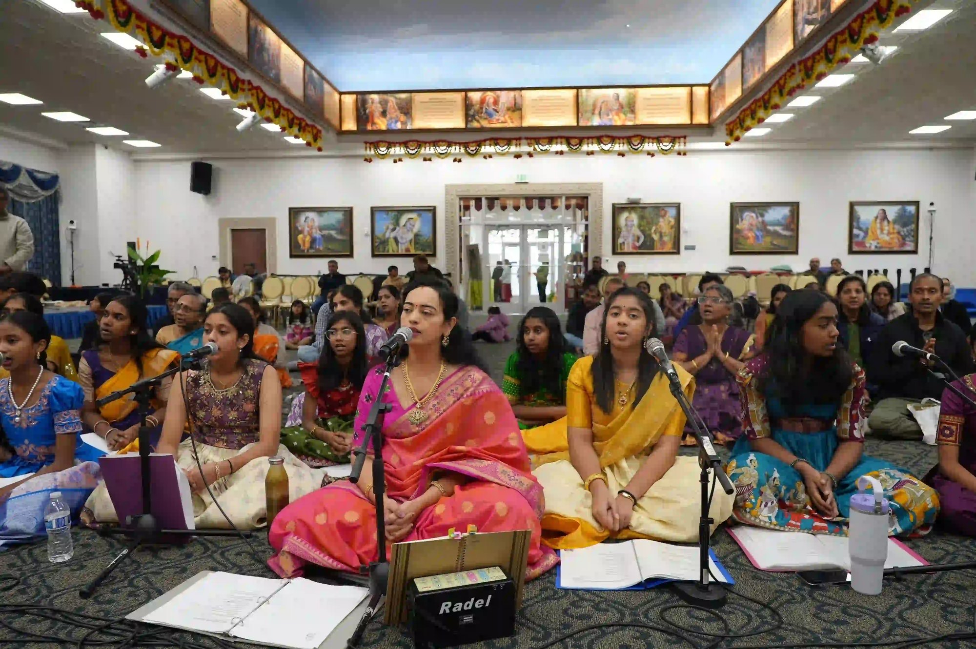 Women in traditional Indian attire seated on the floor performing devotional music inside a temple hall.