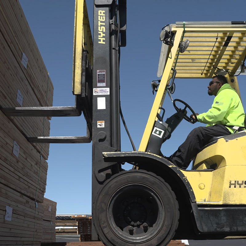 Worker in a high-visibility jacket operating a yellow forklift near stacked wooden planks outdoors.