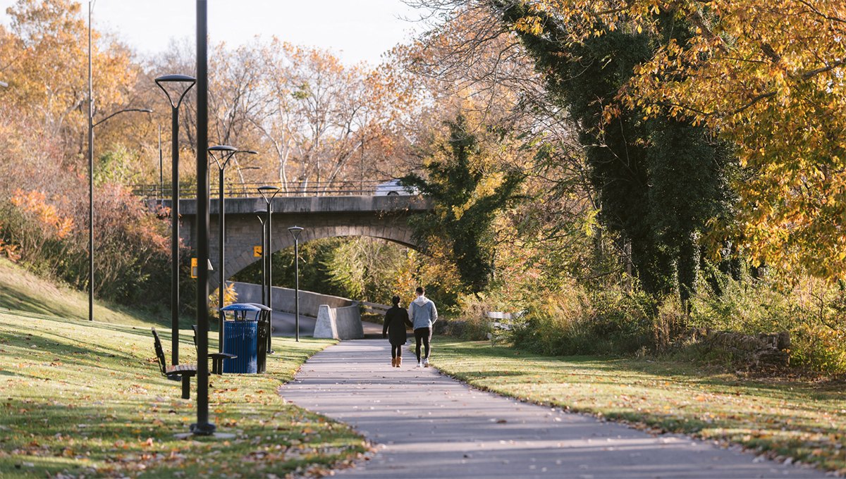 Two people walk side by side on a paved park path lined with autumn-colored trees.