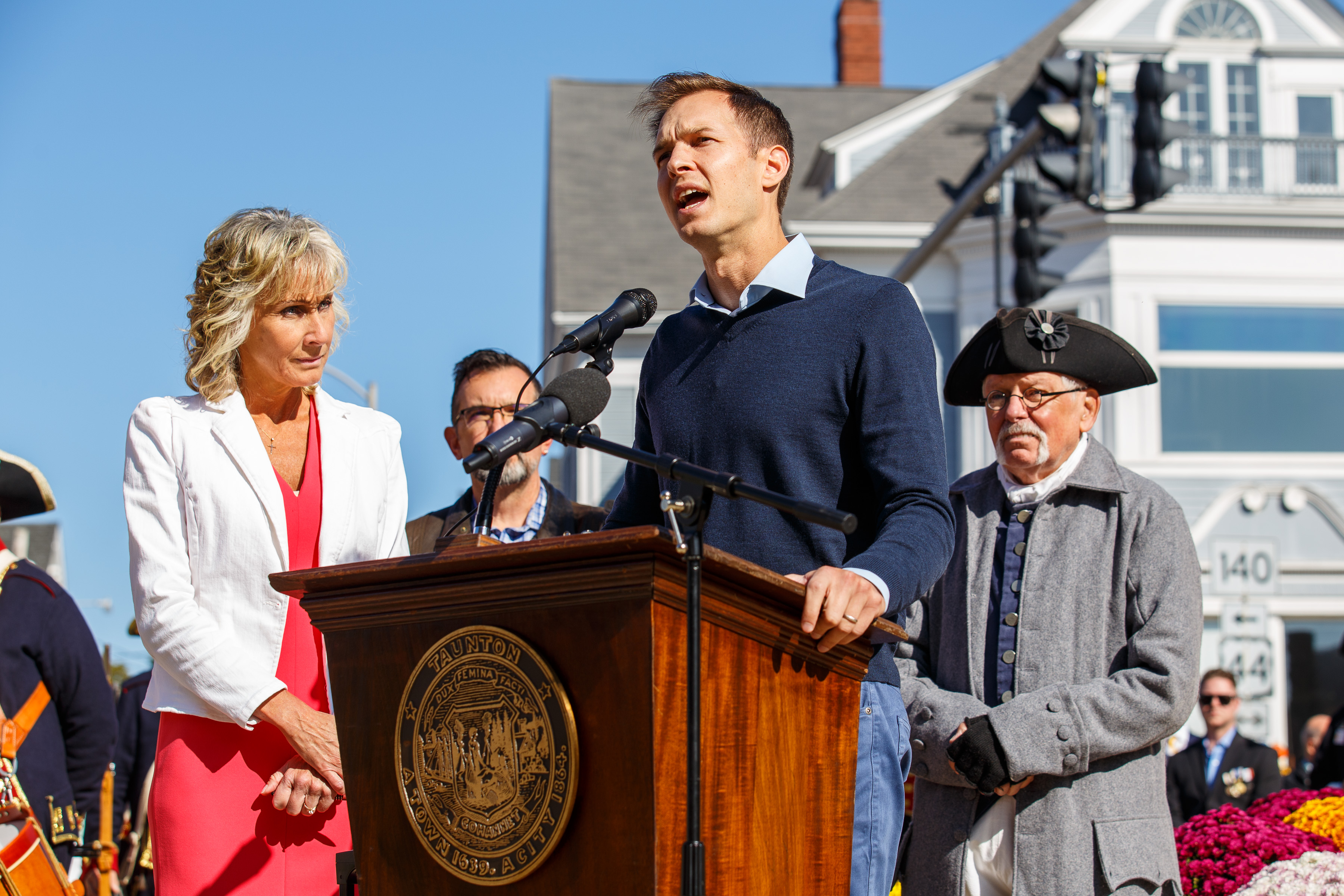 A man speaks at a wooden podium at an outdoor public event.
