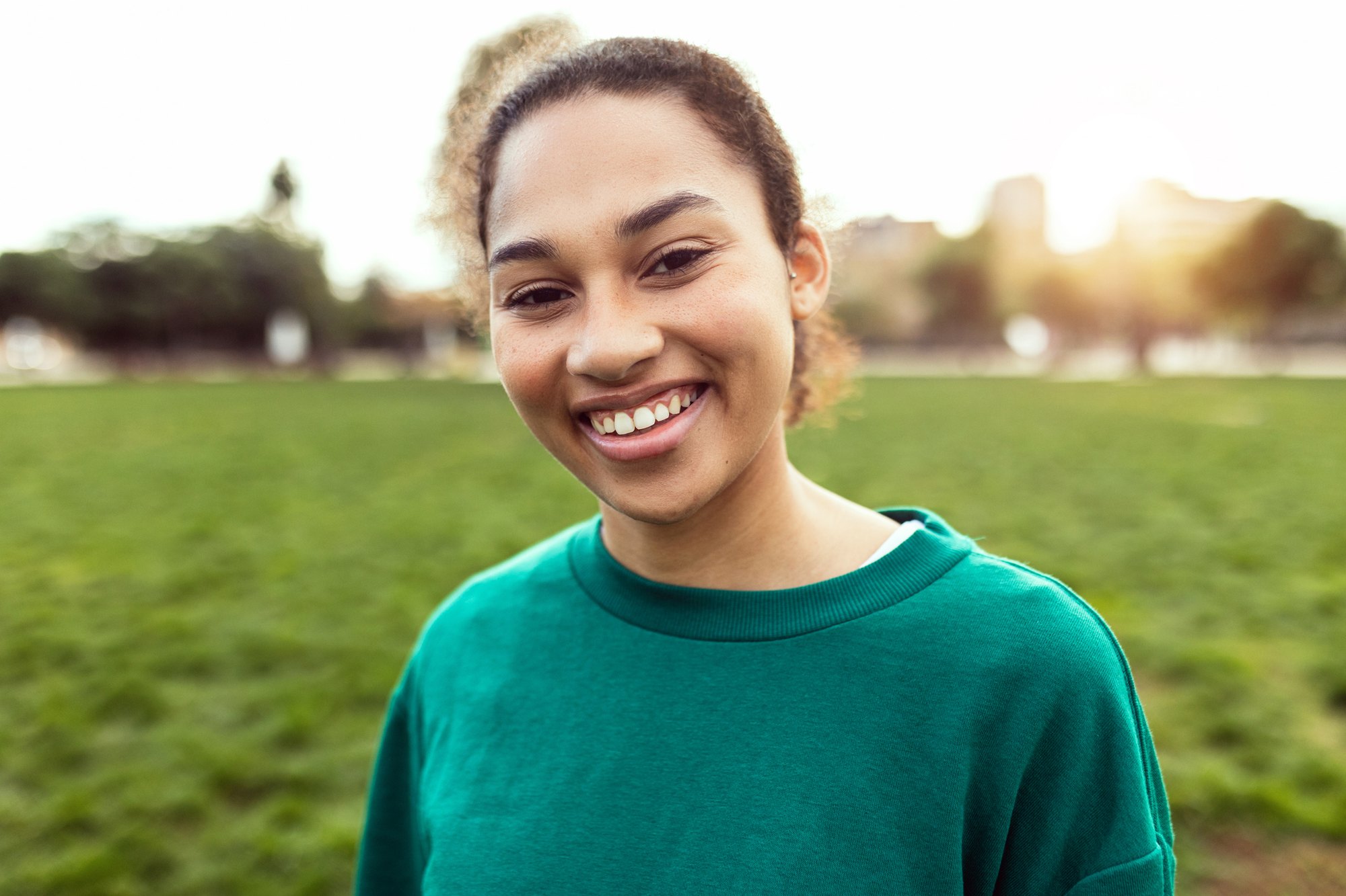 A smiling young woman in a teal sweatshirt standing outdoors in front of trees.
