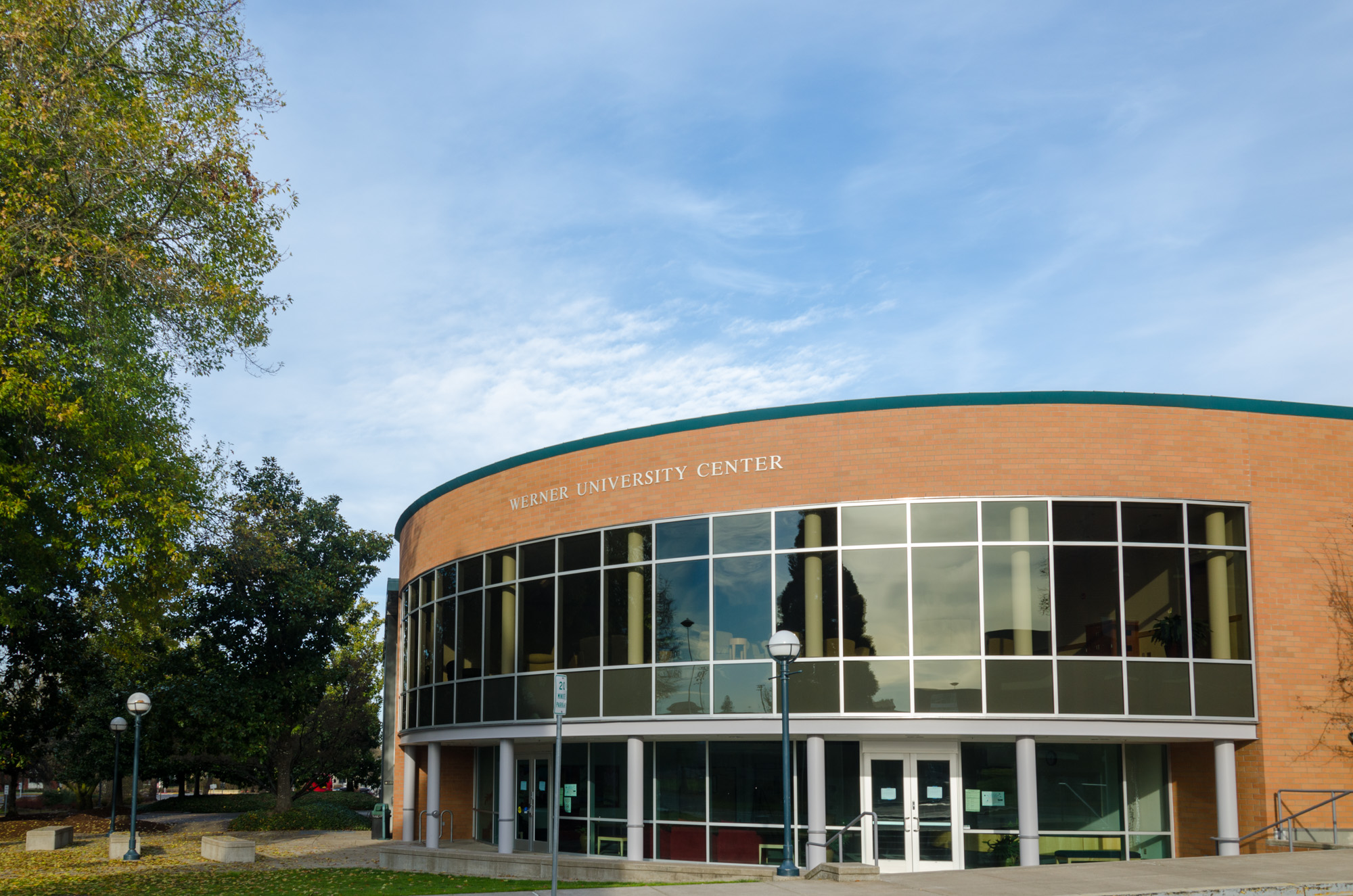 Curved brick building with large glass windows on a university campus under a partly cloudy sky.