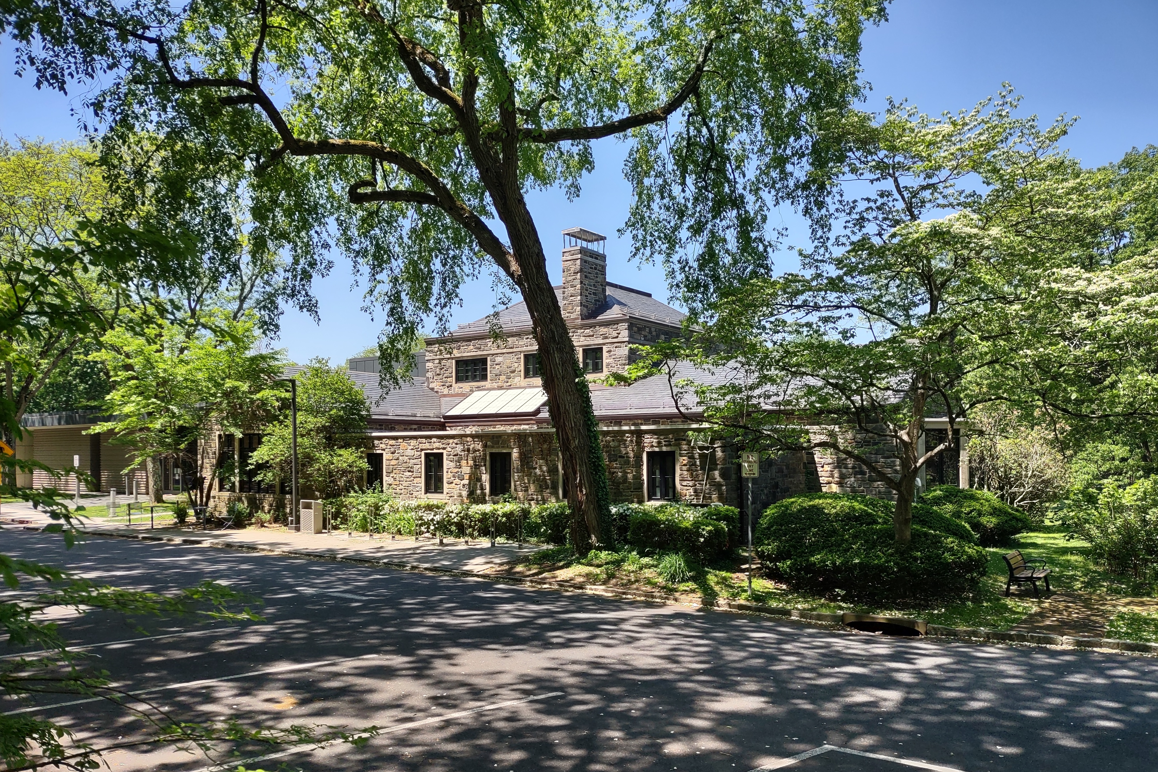 Stone library building with slate roof, partially framed by large mature trees on a sunny day.