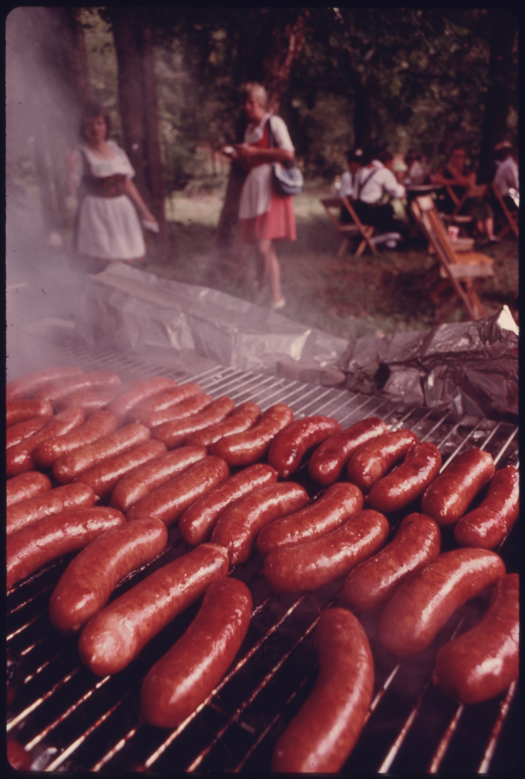 Sausages cooking on a large outdoor charcoal grill at a community festival.
