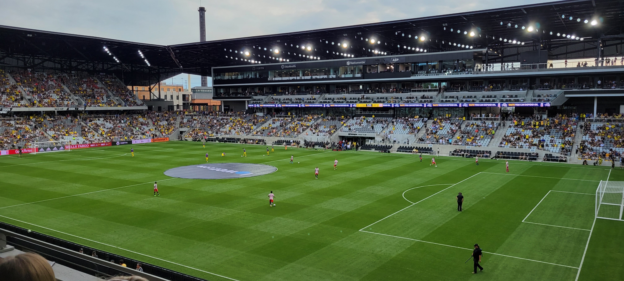 Wide-angle interior view of a soccer stadium with a full crowd and players on a green pitch under floodlights.