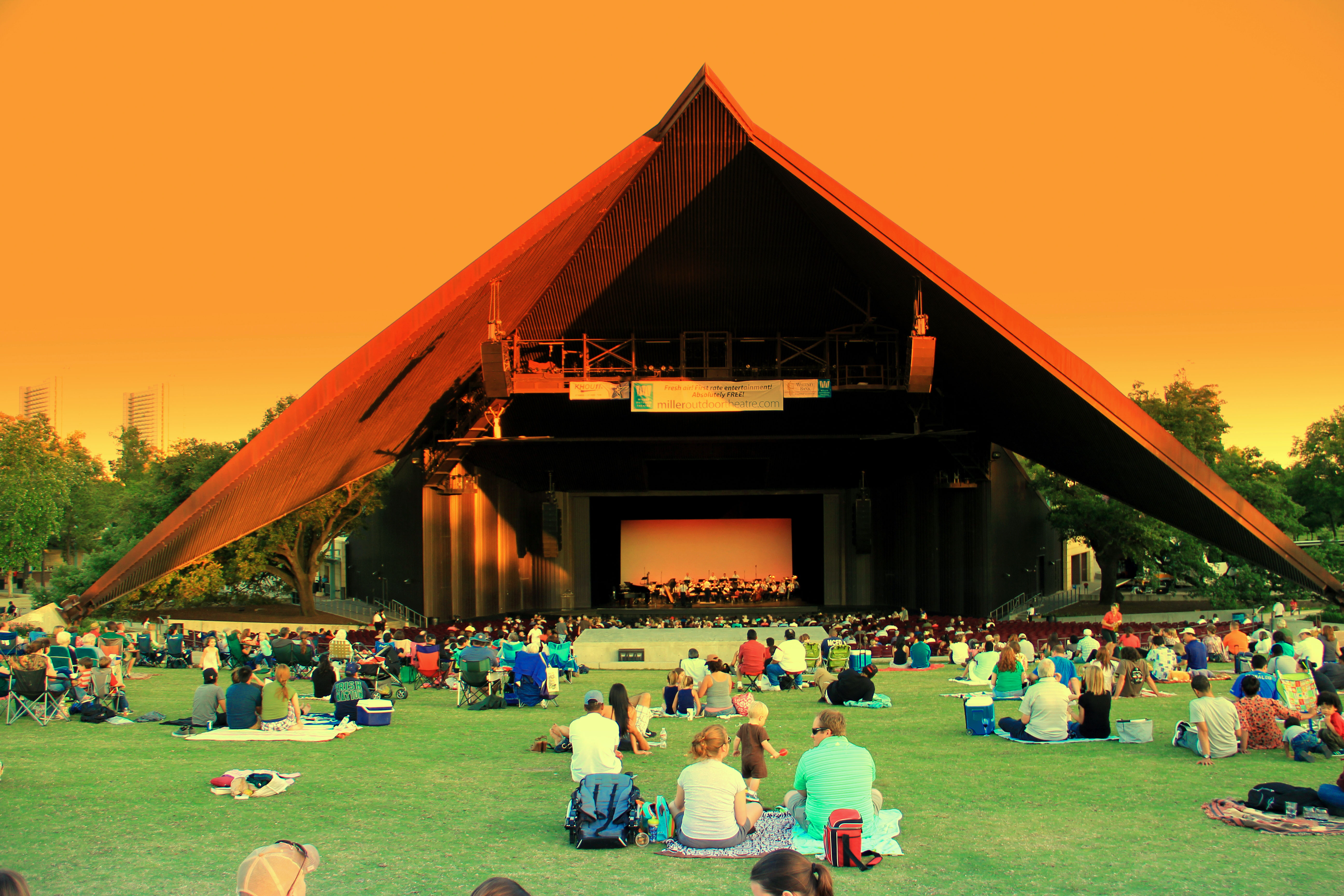 A large crowd sits on a grassy lawn facing an illuminated outdoor stage at dusk.