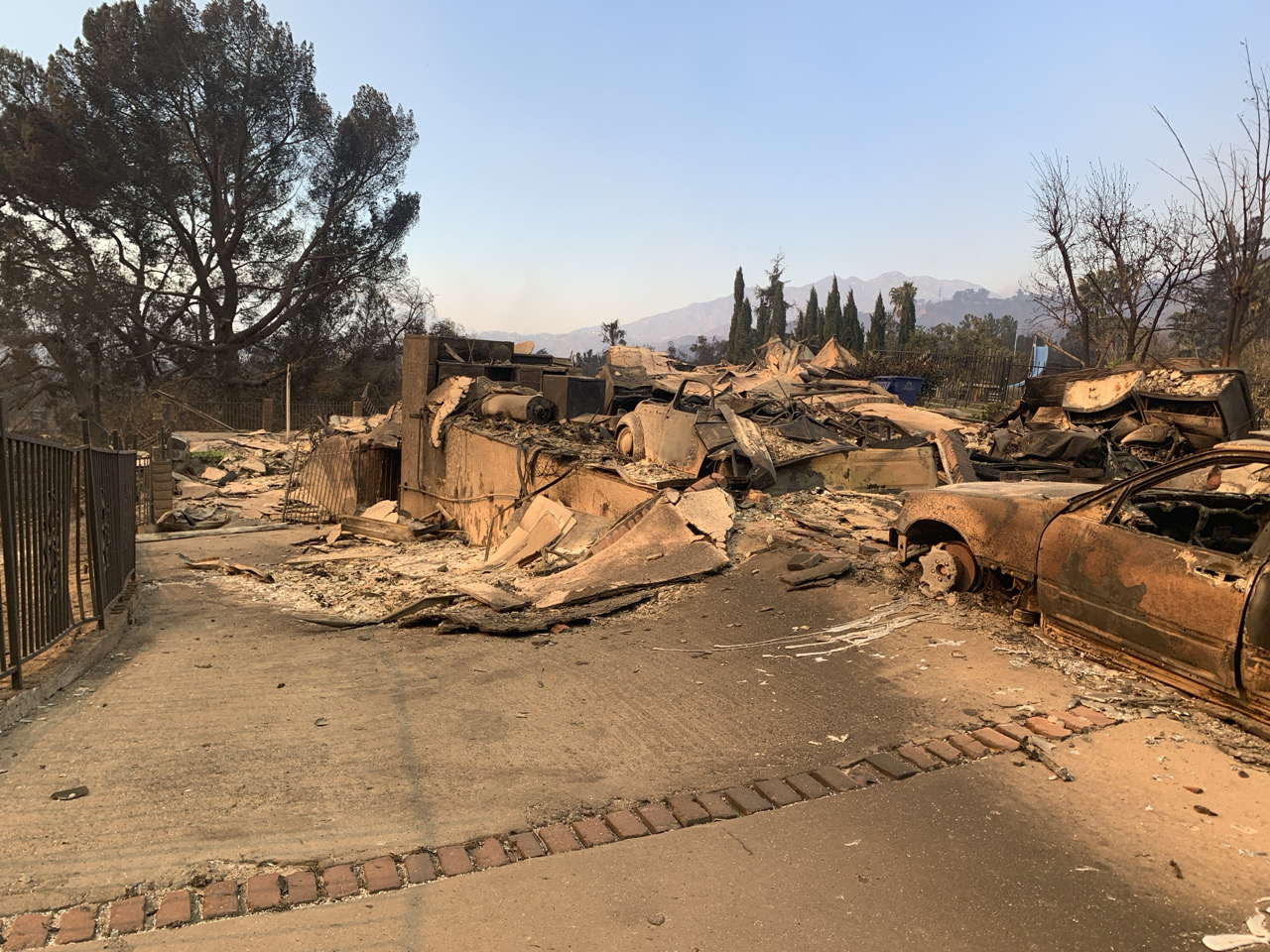 Burned-out home reduced to rubble with a charred vehicle on a residential street.