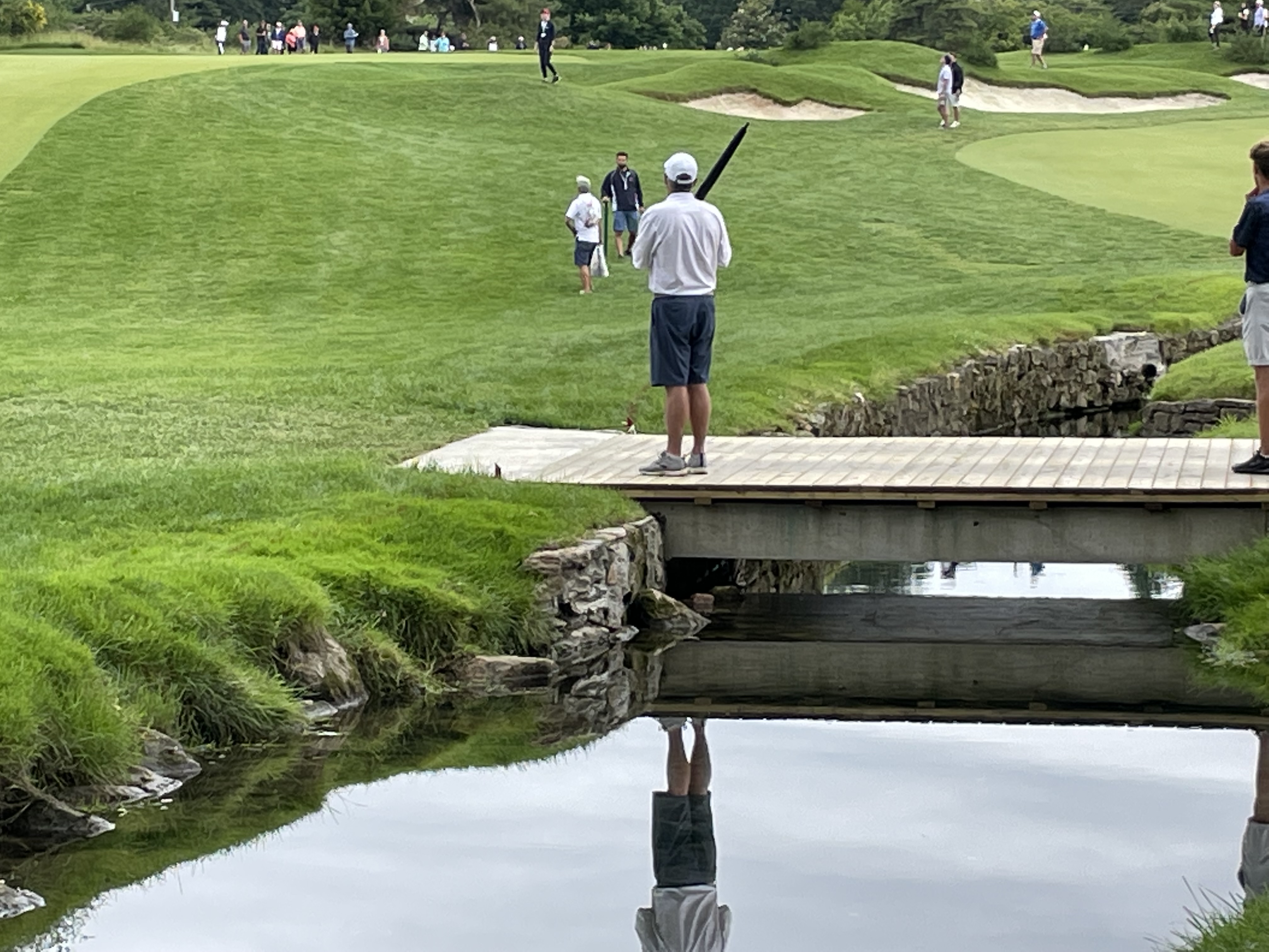Golfer on a wooden footbridge over a water hazard, with bunkers and spectators visible behind.