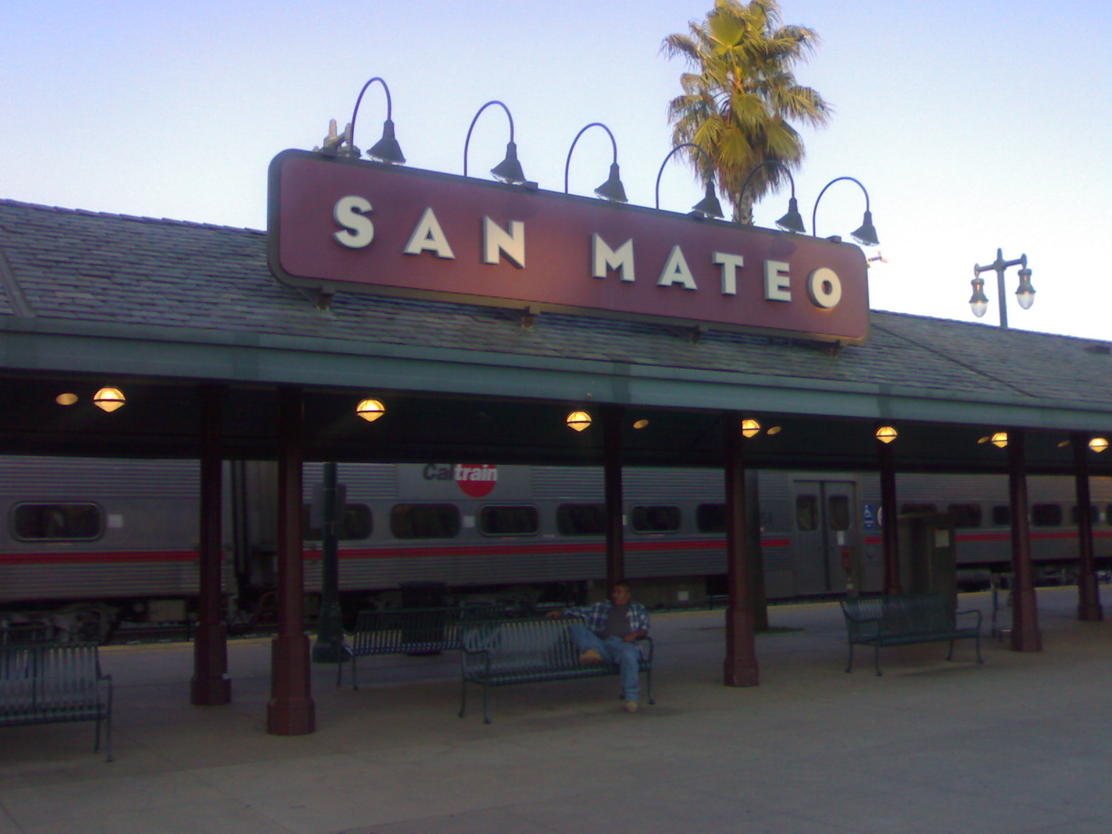 Caltrain station platform at dusk with illuminated sign and a train visible behind it.