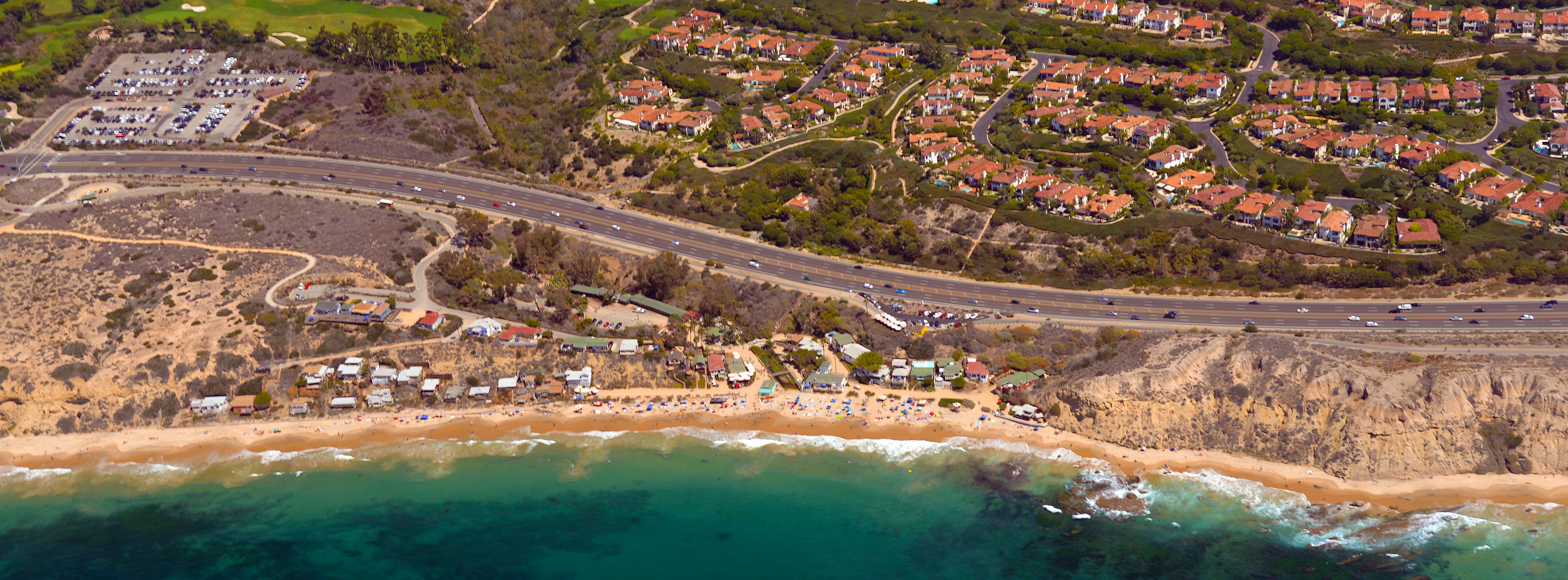 Aerial view of a sandy beach, coastal highway, and hillside neighborhood with red-tile roofs.