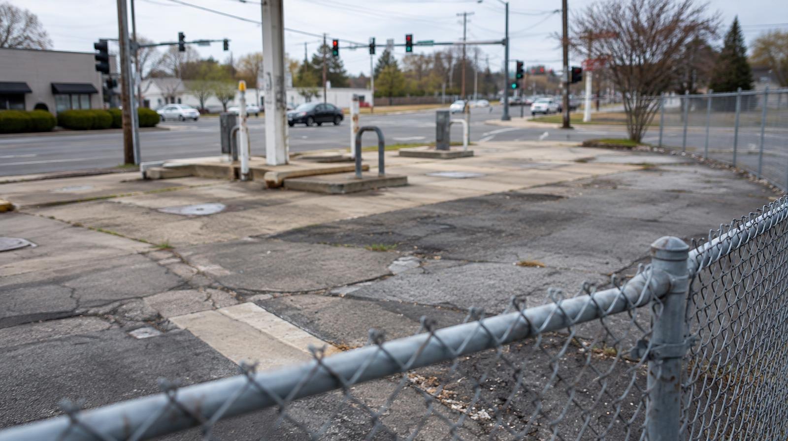 Vacant fenced corner lot with cracked asphalt at a suburban street intersection.