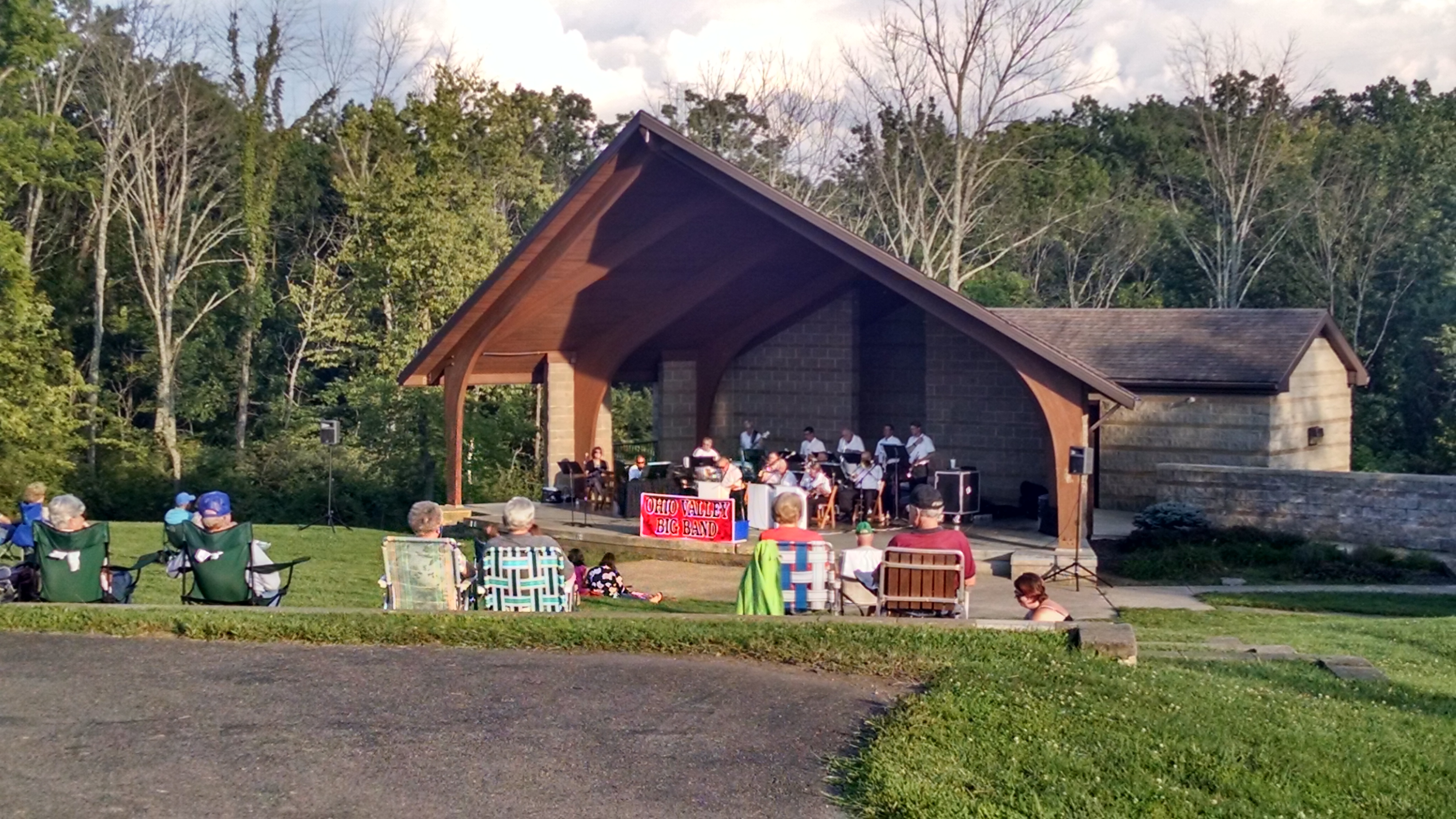 Outdoor amphitheater with peaked wooden roof and stone walls at a park, with musicians performing and audience in lawn chairs.