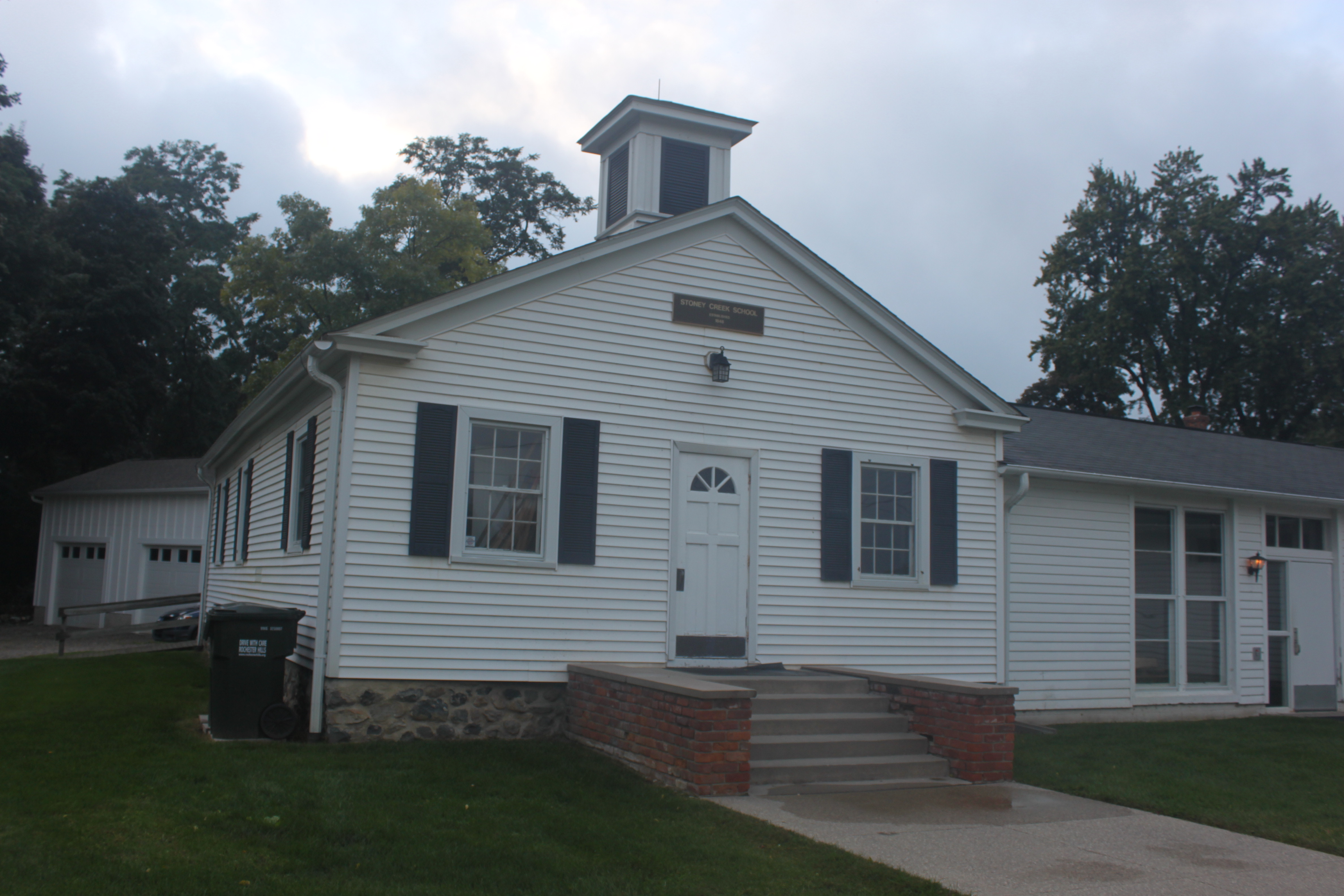 White clapboard schoolhouse with black shutters and a cupola surrounded by trees.