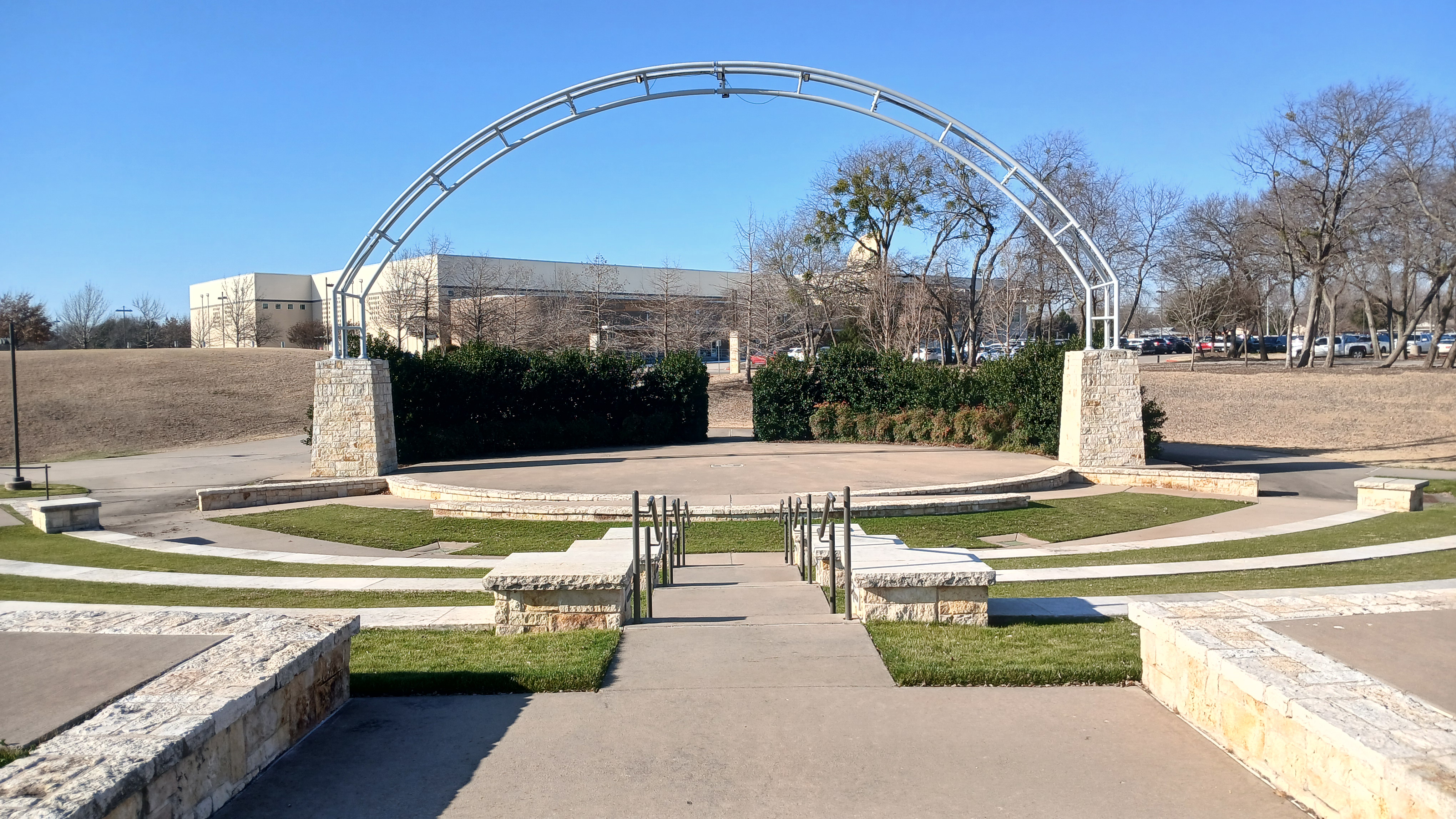 Outdoor amphitheater with a large metal arch and stone pillars under a clear blue sky.