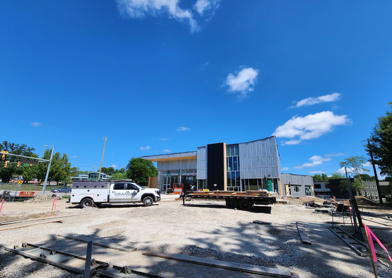 Exterior of Ann Arbor's new Fire Station 4 under construction, showing the building's curved metal-paneled facade and glass entrance, with a Granger construction truck, a flatbed trailer loaded with lumber, and a gravel lot in the foreground under a clear blue sky.