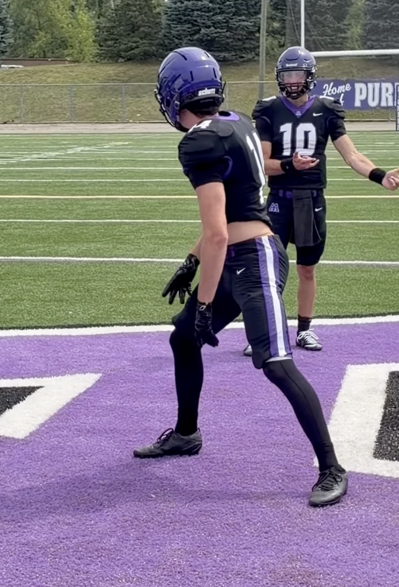 Ann Arbor Pioneer football players in purple and black uniforms run a drill during practice on the turf field.