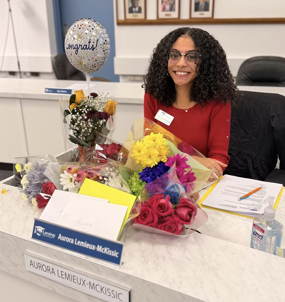 Aurora Lemieux-McKissic smiles behind her Lansing school board nameplate, surrounded by flower bouquets and a congratulatory balloon.