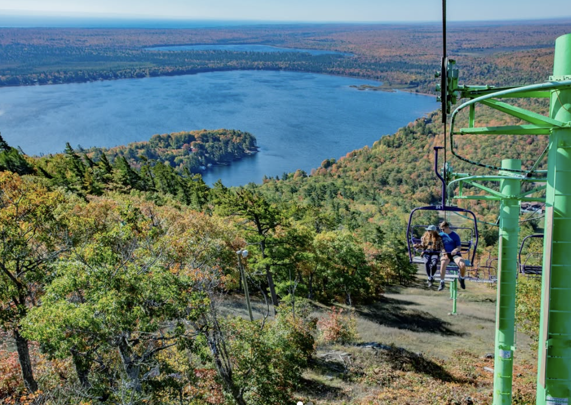 Two people ride a chairlift above a wooded hillside with autumn foliage overlooking a blue lake.