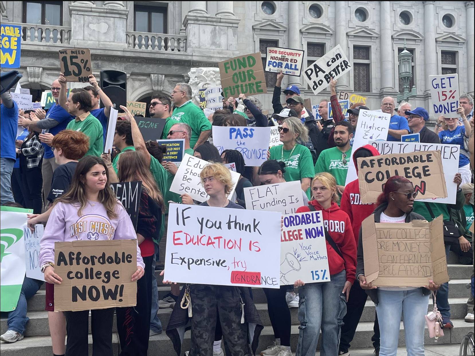 A crowd of protesters on government building steps holding signs about affordable college funding.