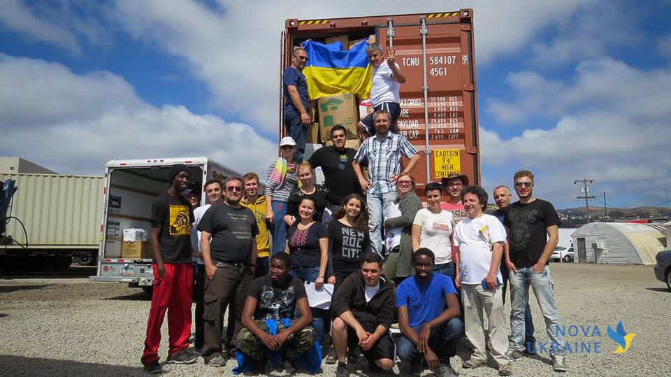 Group of about 20 volunteers posing in front of an open shipping container with a Ukrainian flag.