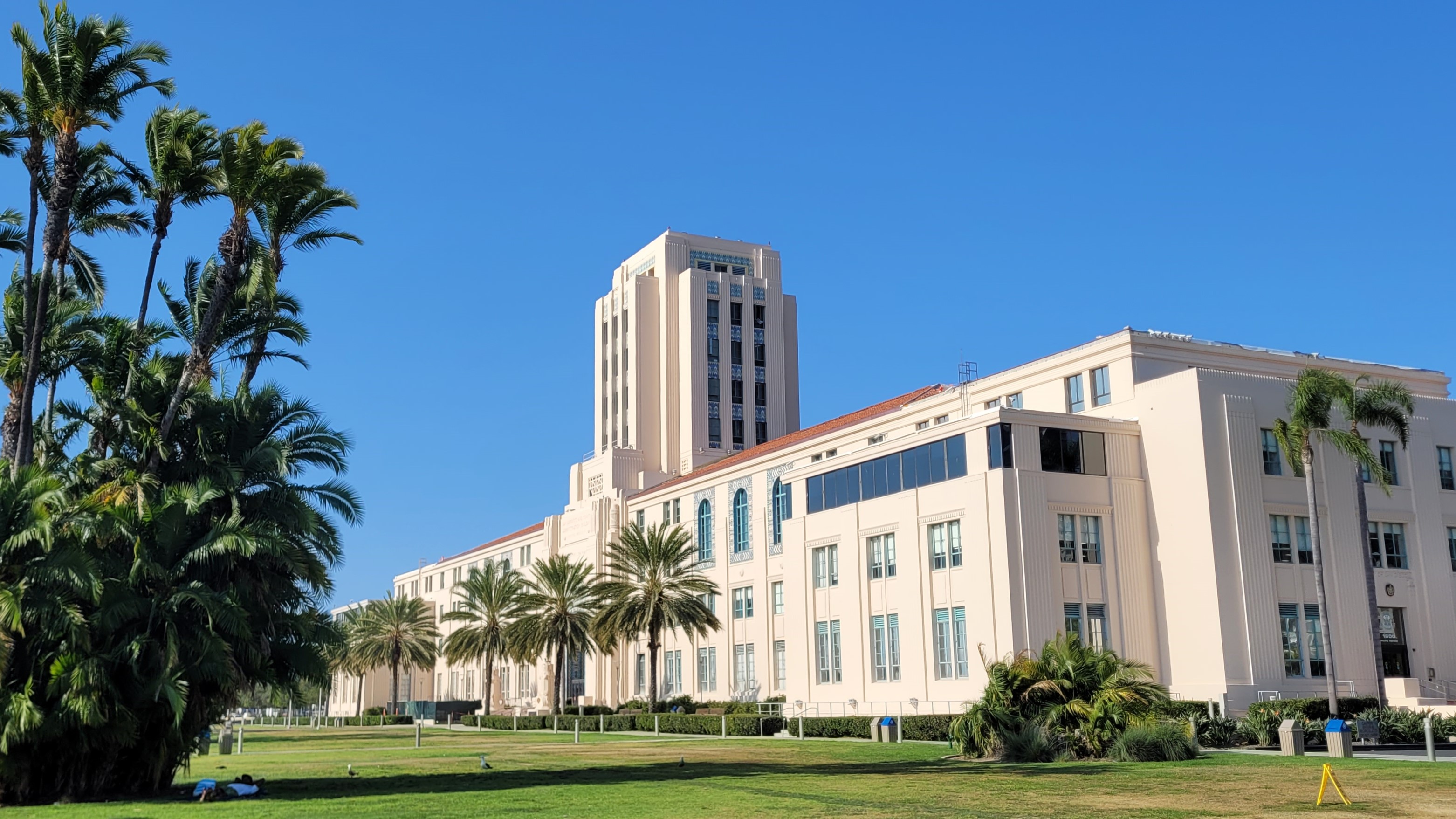 Art Deco government building with central tower, palm trees, and green lawn under blue sky.