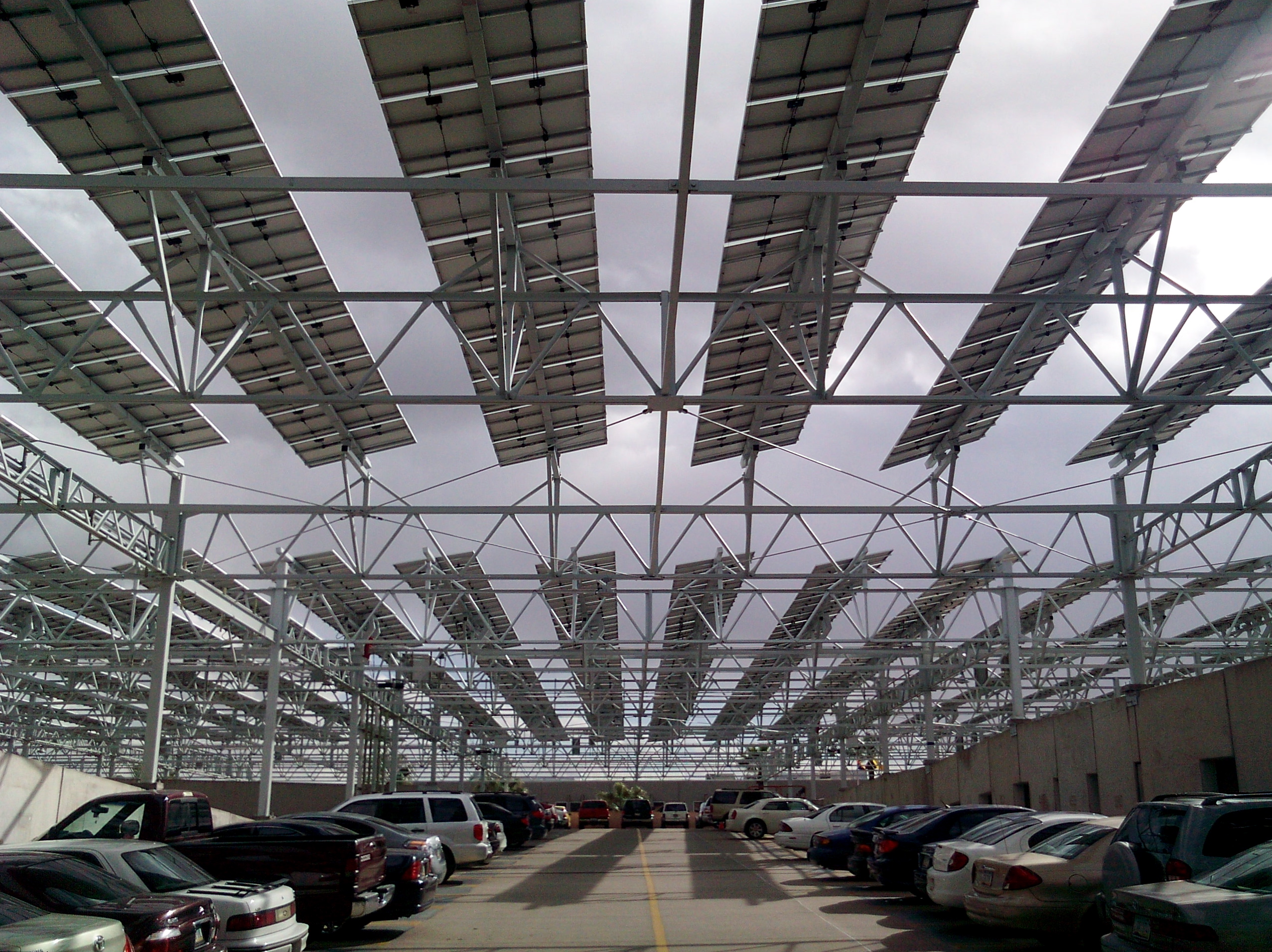 Solar photovoltaic panels mounted on a steel canopy over a parking lot full of cars.