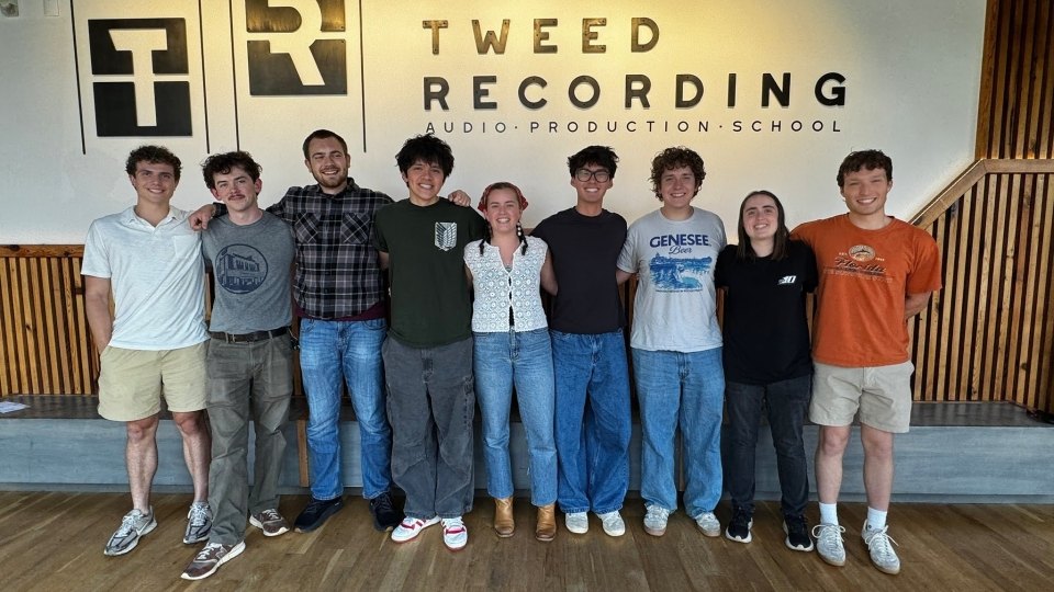 Nine young adults stand in a line with arms around each other inside a modern recording school lobby.