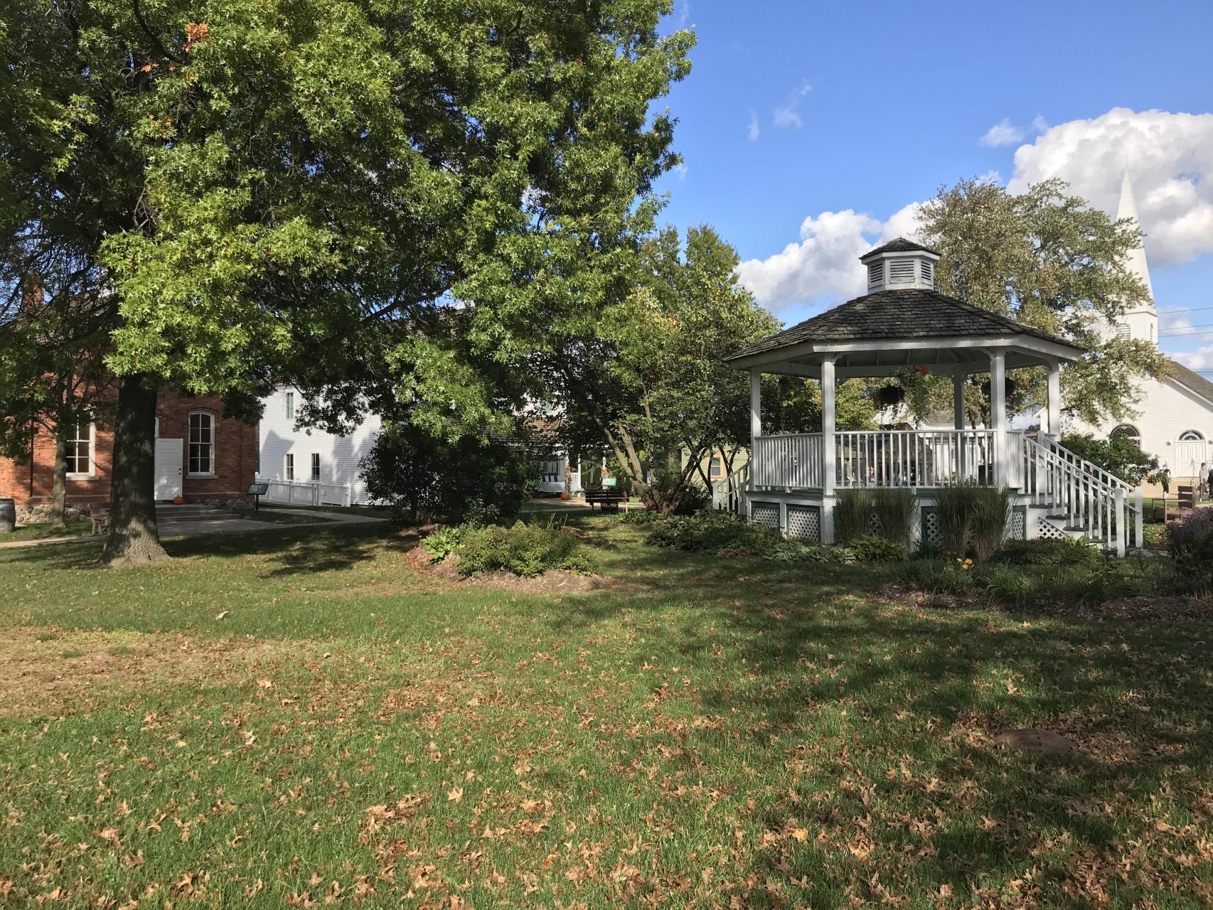 White gazebo with historic buildings and mature trees on a green lawn.