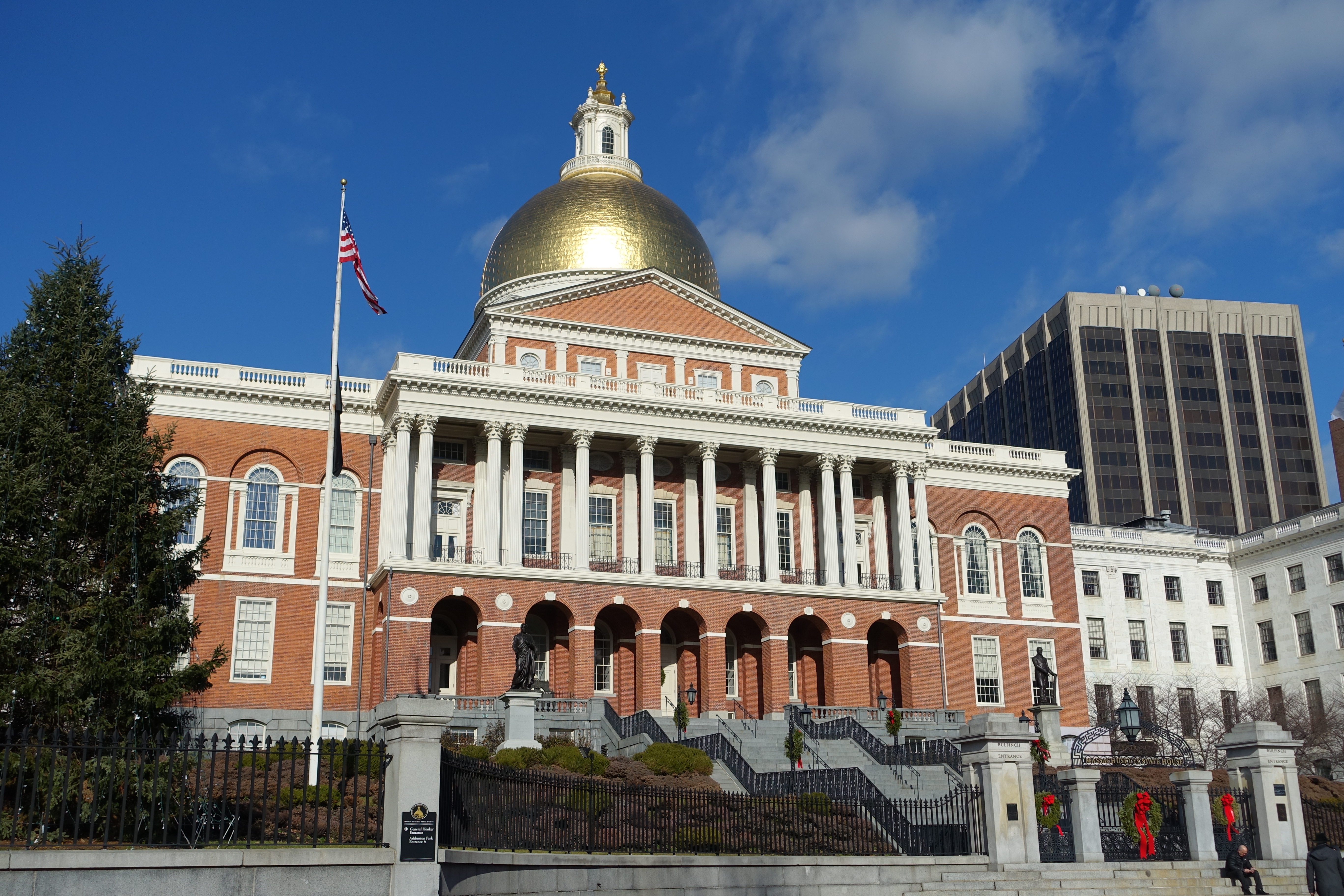 Massachusetts State House exterior with gold dome, white columns, and an American flag on a clear day.