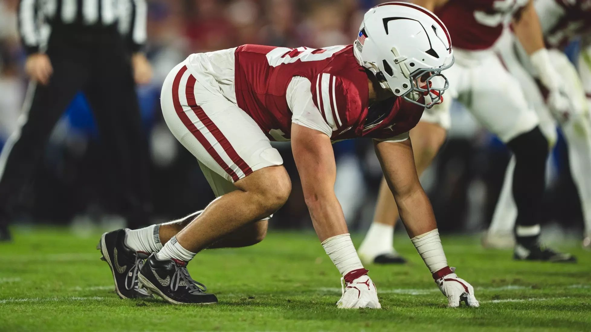 A football player in a red and white uniform crouches in a defensive lineman's stance on a grass field.