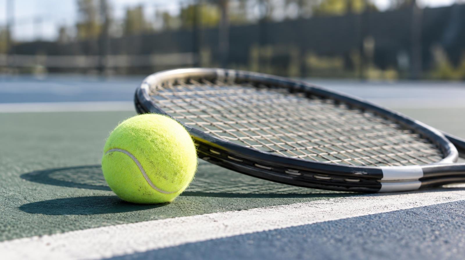 A tennis racket and yellow tennis ball resting on a hard court surface near the baseline.