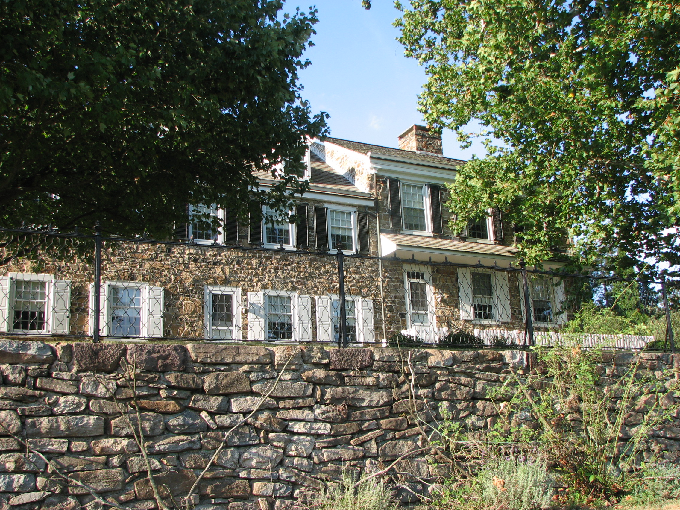 Stone two-story manor house with shuttered windows behind a stone retaining wall.