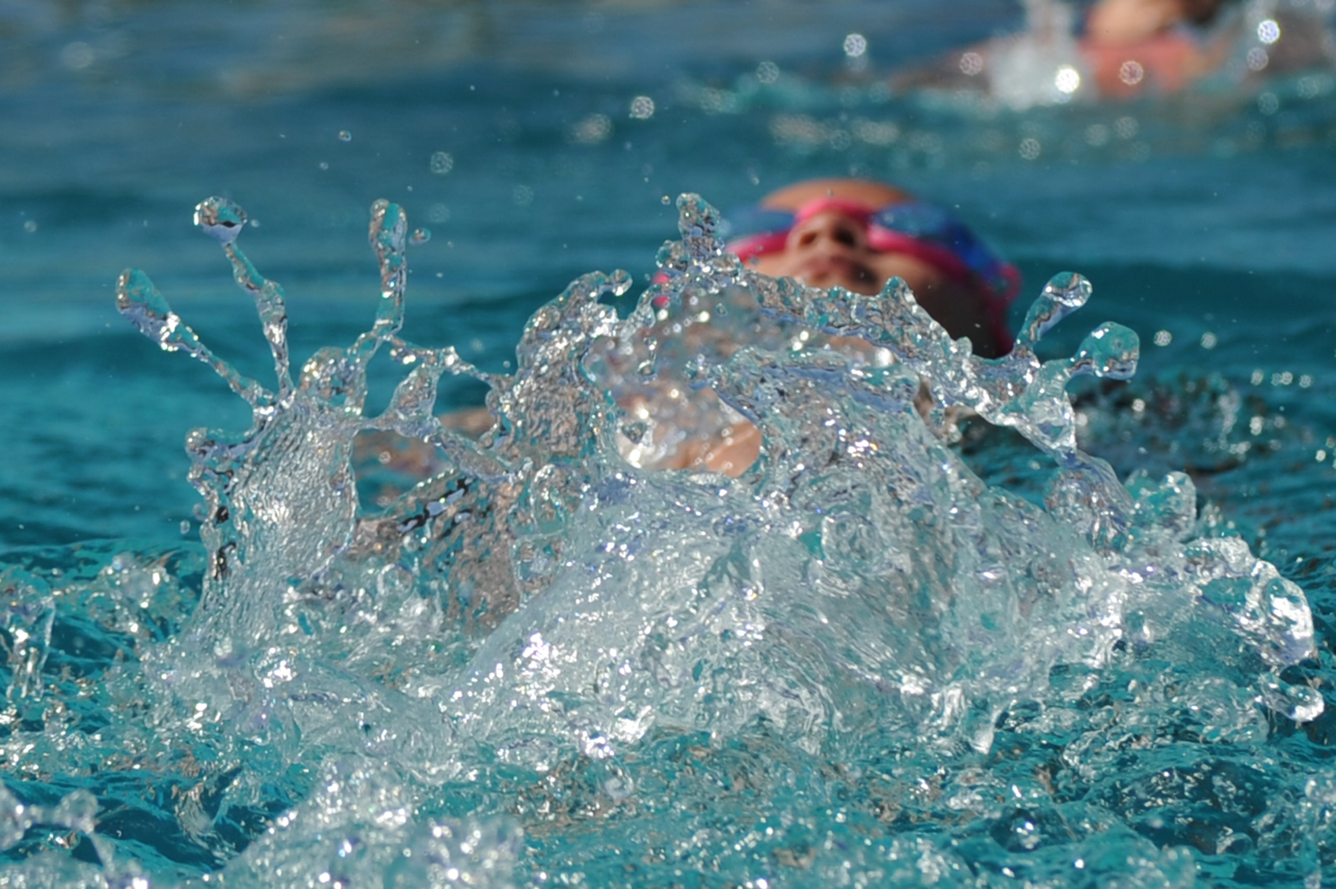 Swimmer in a pink cap doing backstroke with a large water splash in the foreground.
