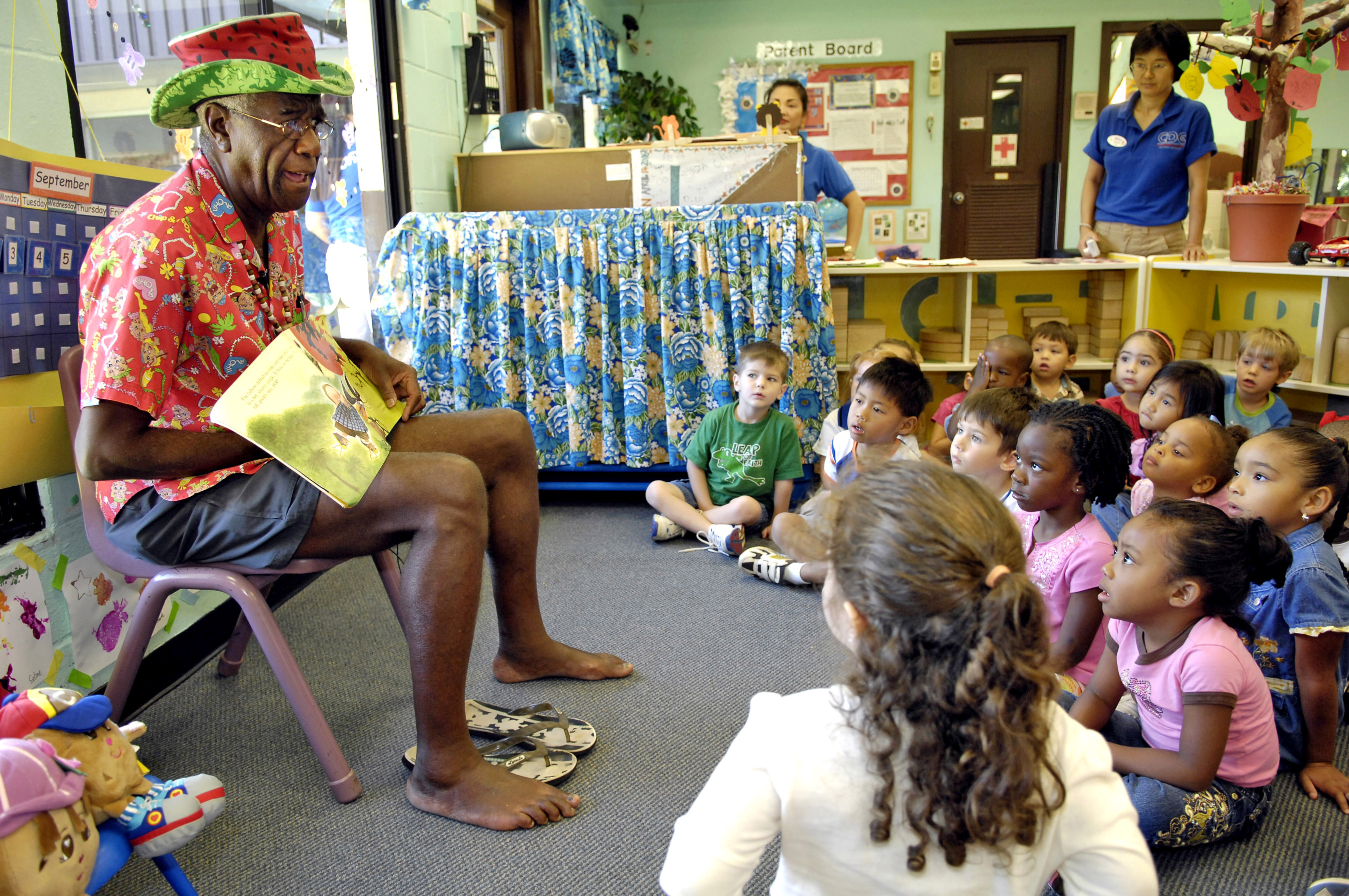 An adult in a colorful shirt reads a picture book to young children on a floor.