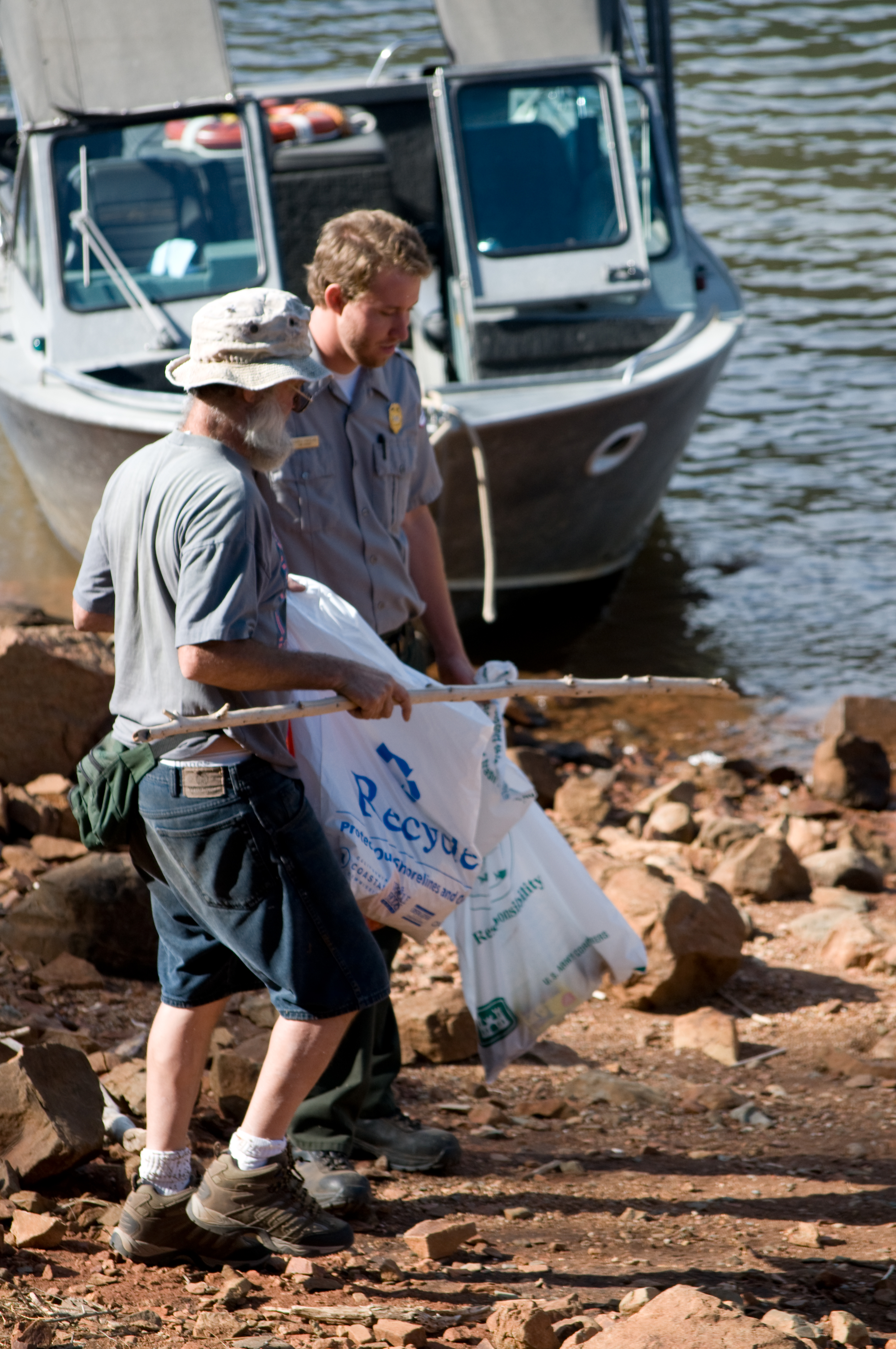 Two people holding a recycling bag picking up litter on a rocky shoreline.