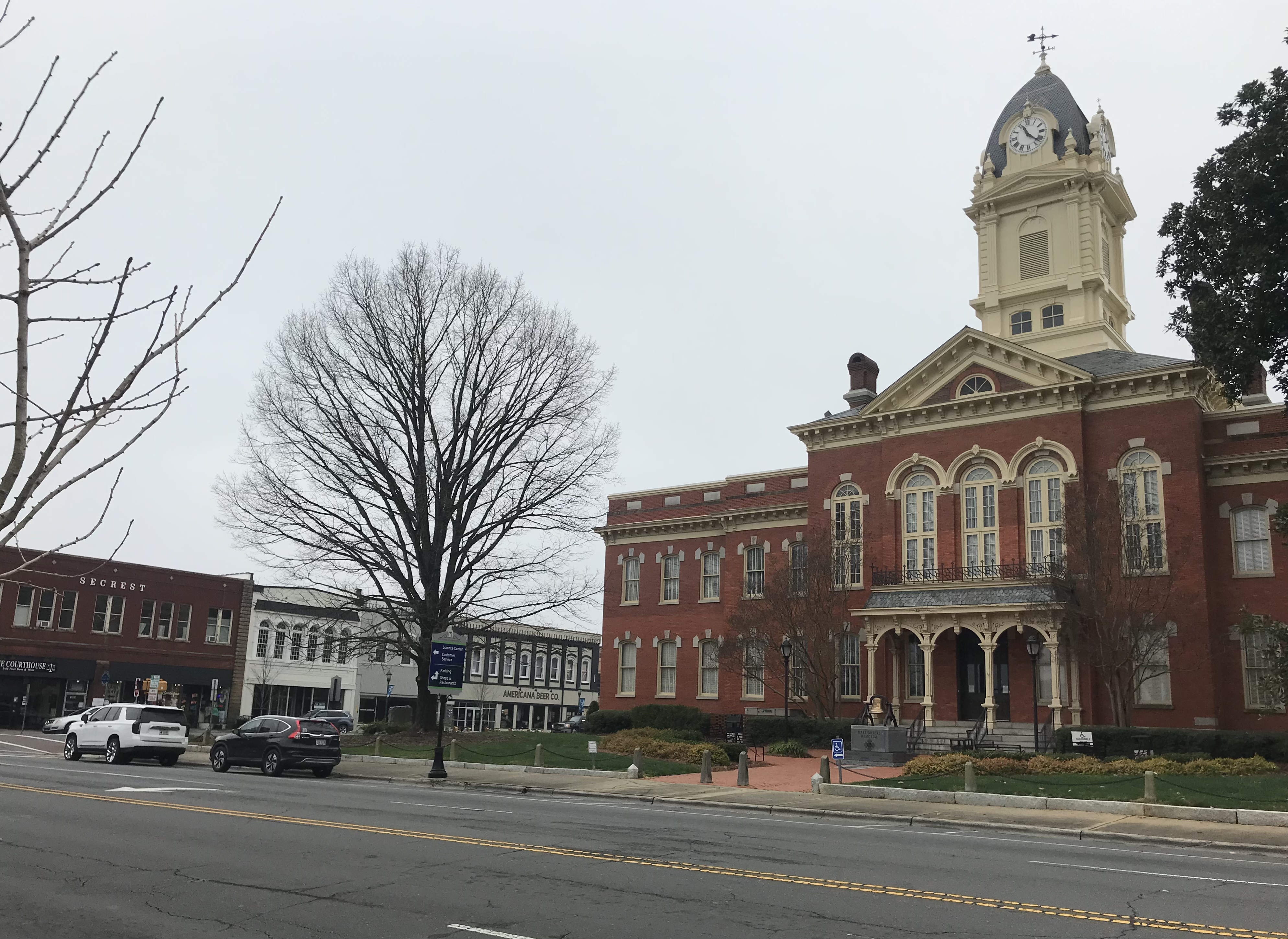 Red-brick courthouse with clock tower flanked by downtown commercial buildings along a street.