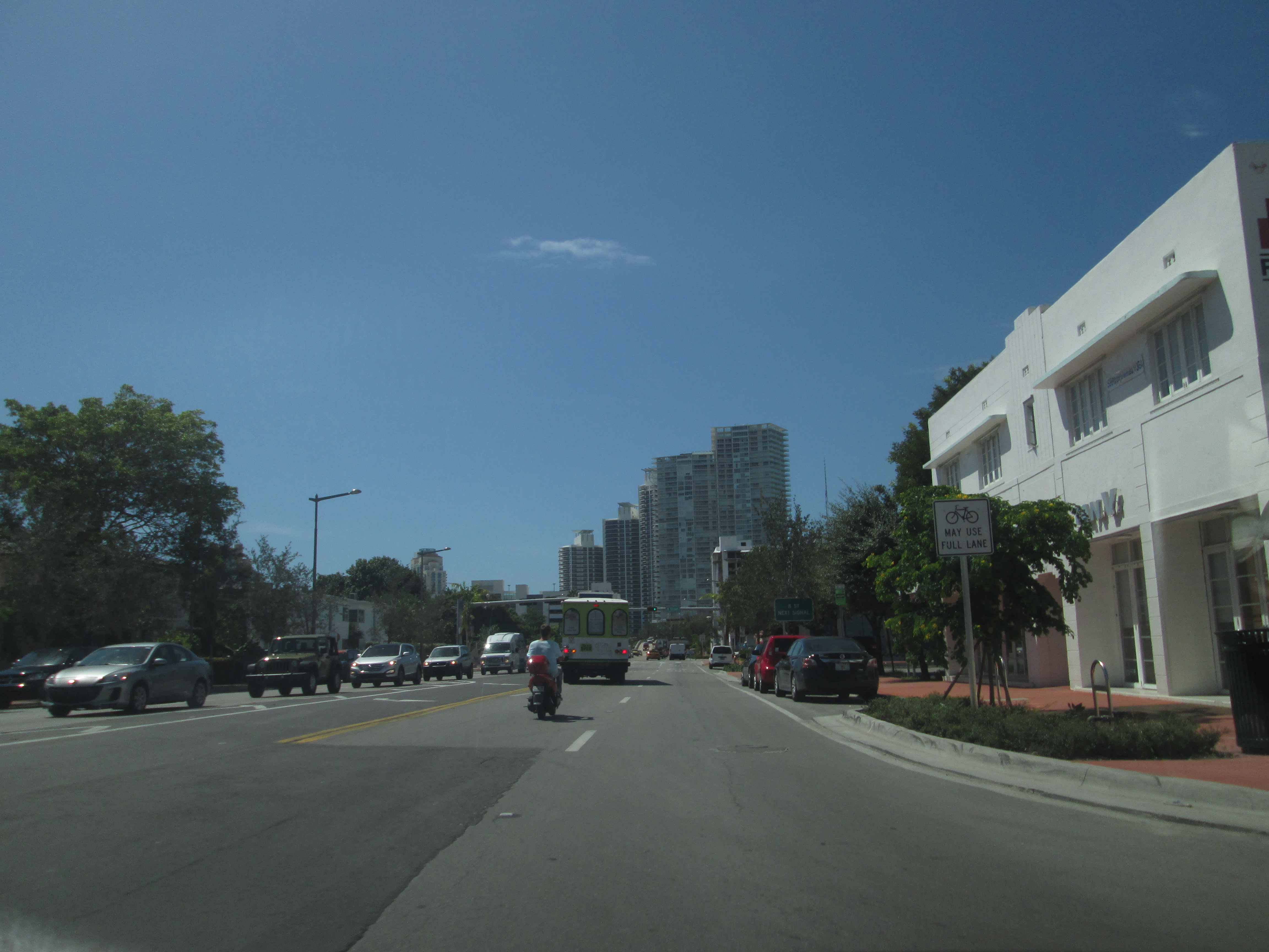Cars and a bus travel north along Alton Road lined with palm trees and a white Art Deco building.