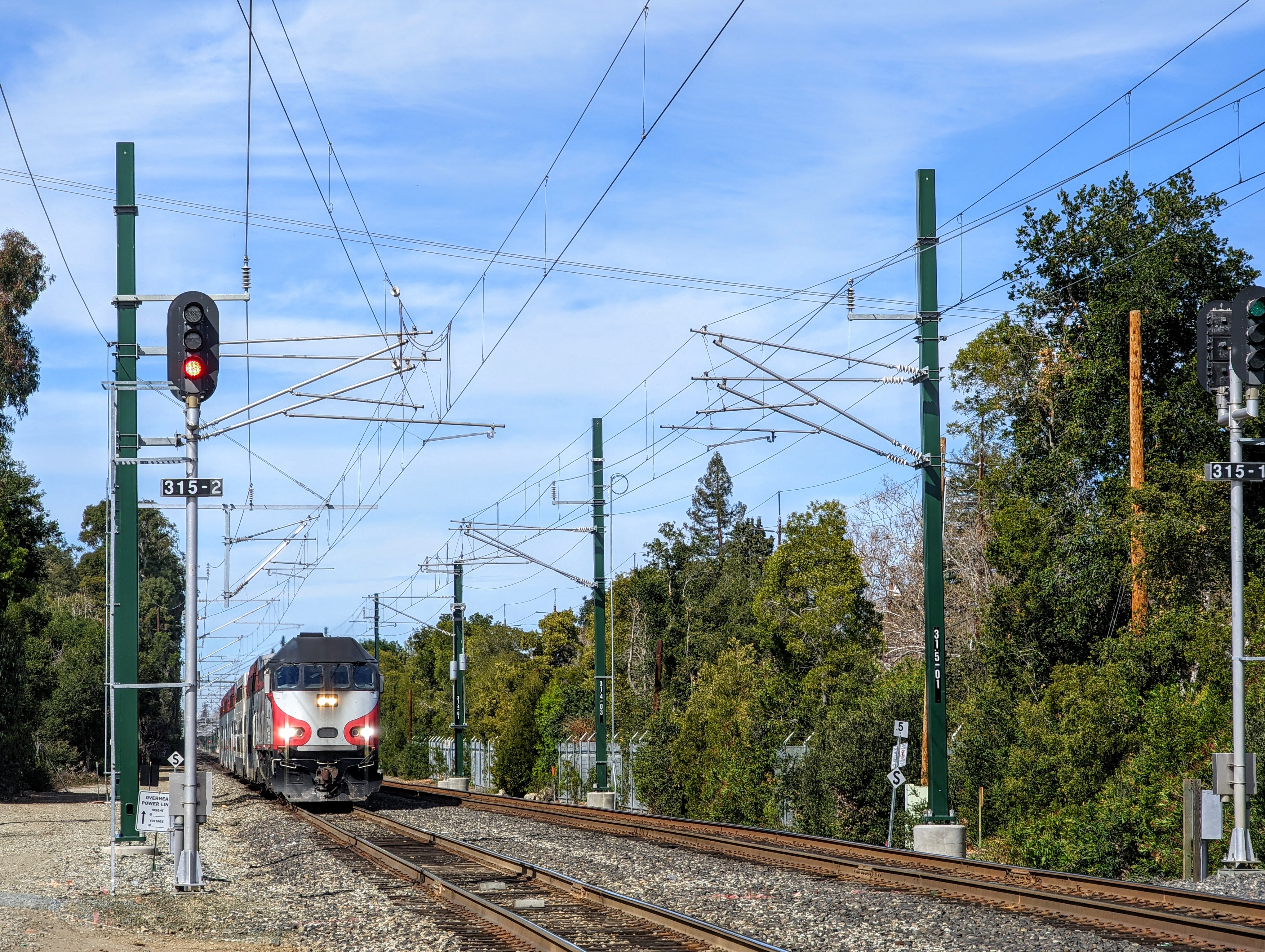 Caltrain train on electrified tracks with overhead catenary wires and signal poles.