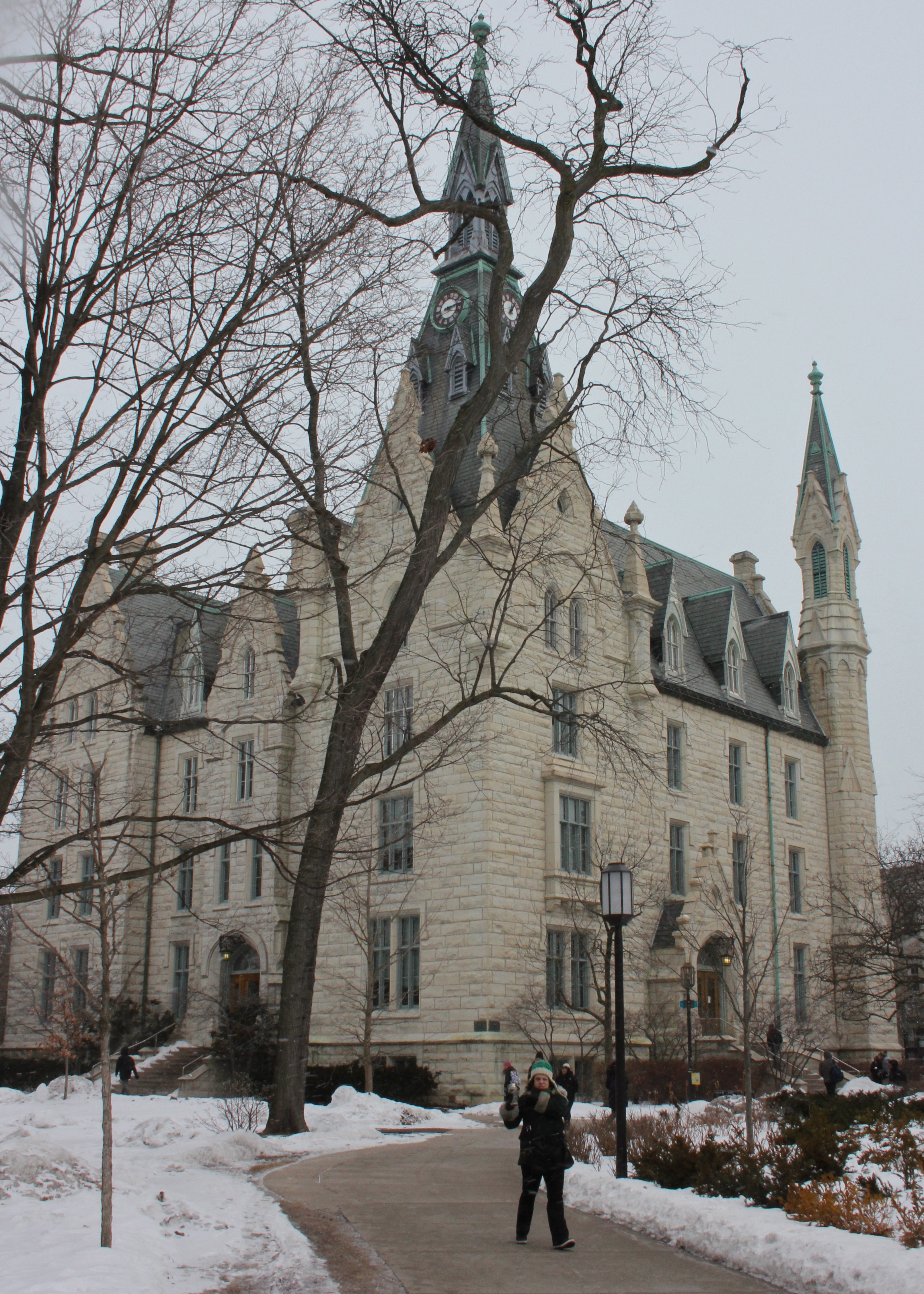 Gothic stone building with clock tower and spires amid snow-covered grounds in winter.