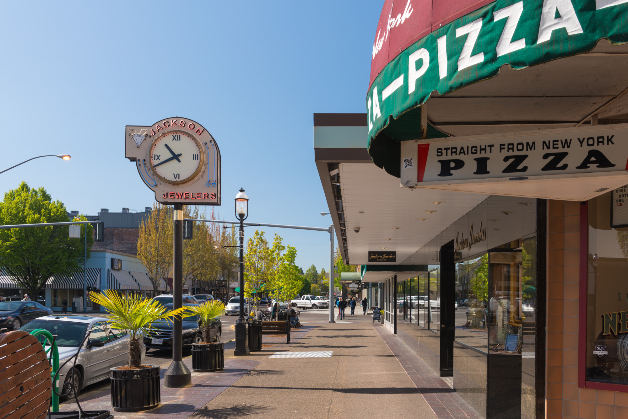 Wide sidewalk along a downtown commercial street with benches, planters, and a clock sign.