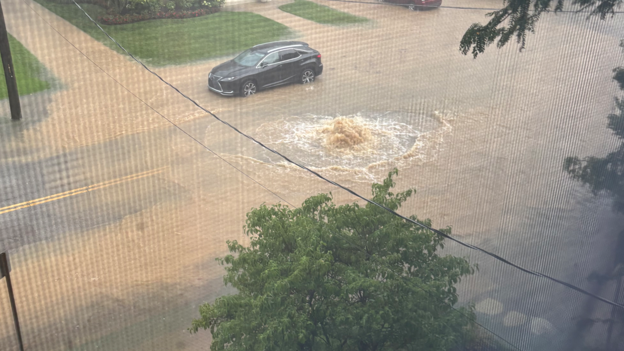 Muddy water gushing upward from a manhole on a heavily flooded street.