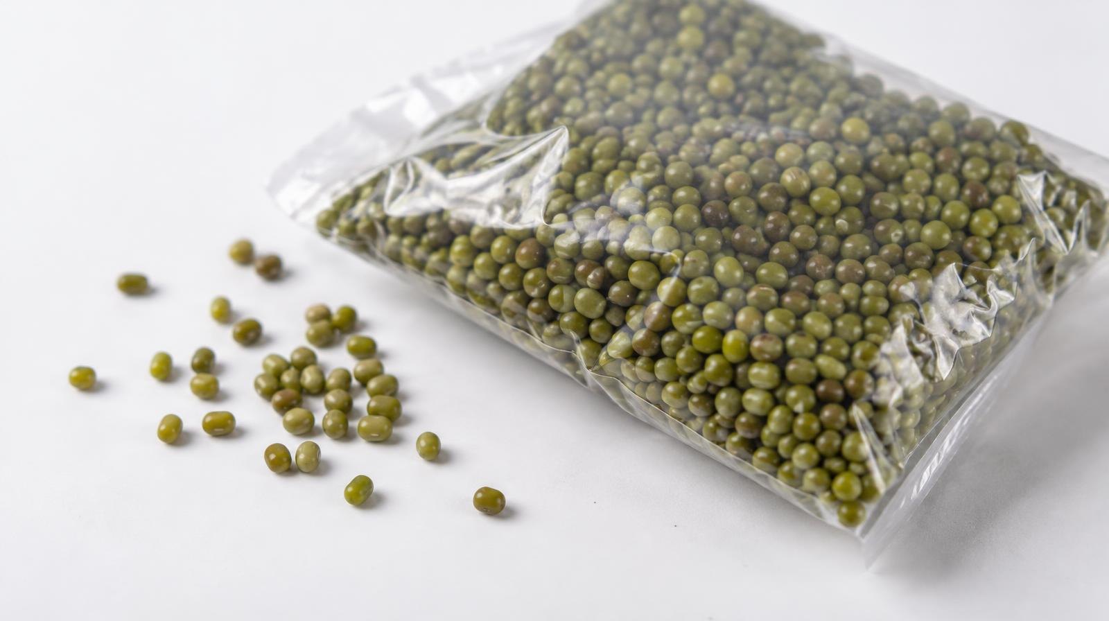 A clear plastic bag of dried green mung beans on a white surface with loose beans beside it.