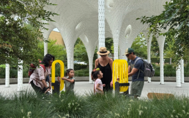 Adults and children gather around yellow arch-shaped sculptures under an outdoor pavilion.