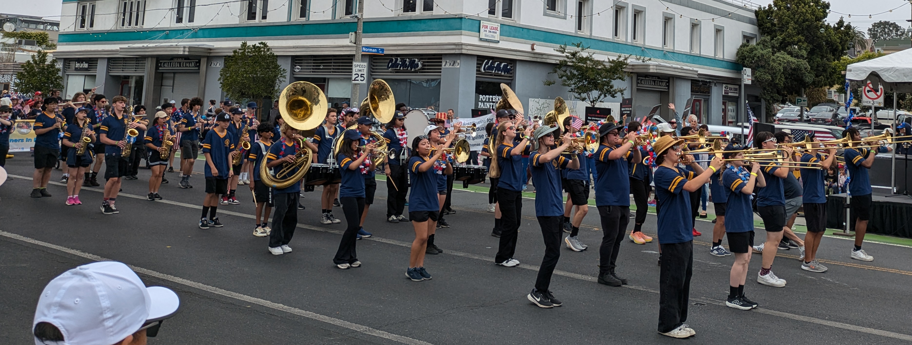 Marching band in dark blue uniforms playing brass instruments during a parade on a commercial street lined with spectators.