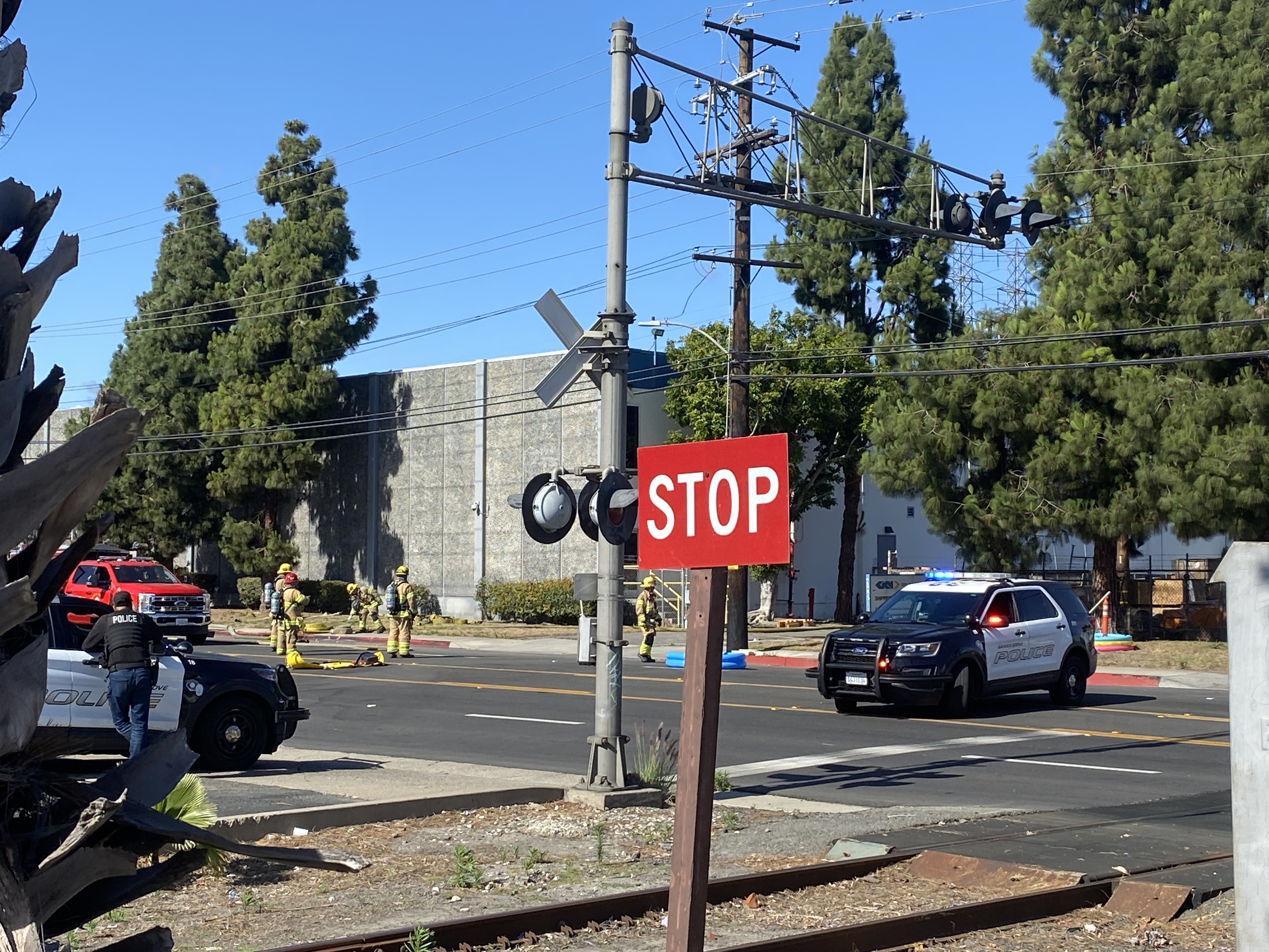 Firefighters in gear and police vehicles near a railroad crossing outside a large industrial building.