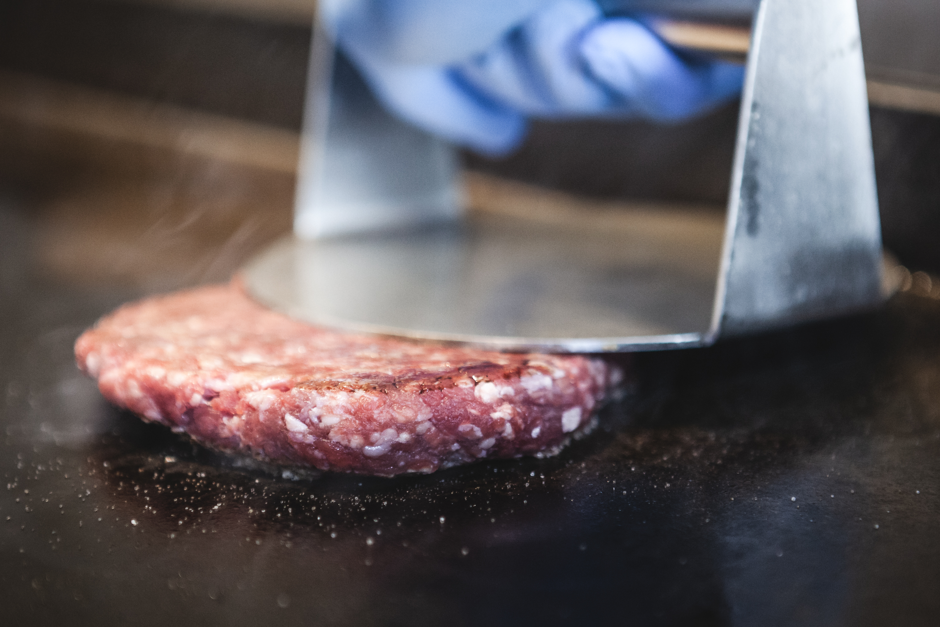 A gloved hand presses a metal spatula onto a beef patty sizzling on a griddle.